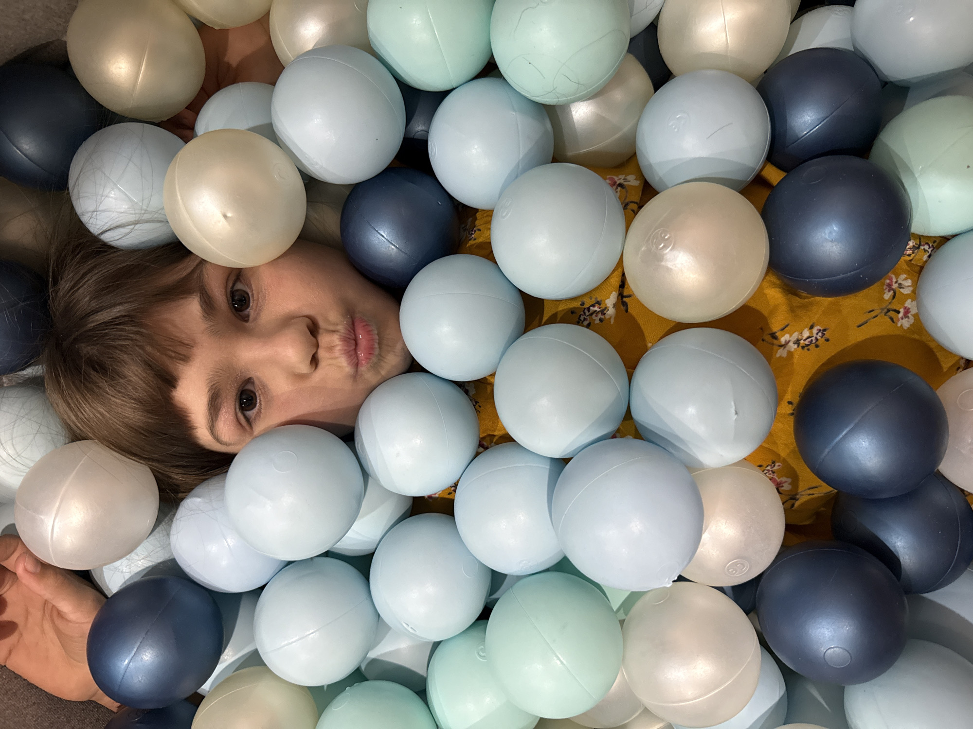 A girl with brown hair and light skin is partially submerged in a pit of multi-colored plastic balls. The balls are varying shades of light blue, white, and dark blue. A patch of yellow fabric with a floral pattern is visible underneath the balls around the girl's face. The location, Rusanovo, Russia, cannot be confirmed from the image alone.