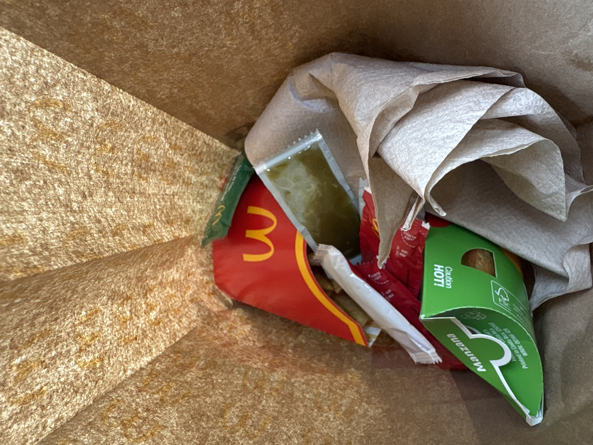 A top-down, close-up shot shows the inside of a tan paper bag containing remnants of a McDonald's meal. A crumpled paper napkin sits atop the contents. Visible are a red McDonald's french fry container, a green "Manzana" pastry box with a "Caution HOT!" warning, a sauce packet, and a partially eaten french fry in a white wrapper. The scene likely takes place where McDonald's is present, potentially in Mexico City, Mexico, indicated by the "Manzana" box.