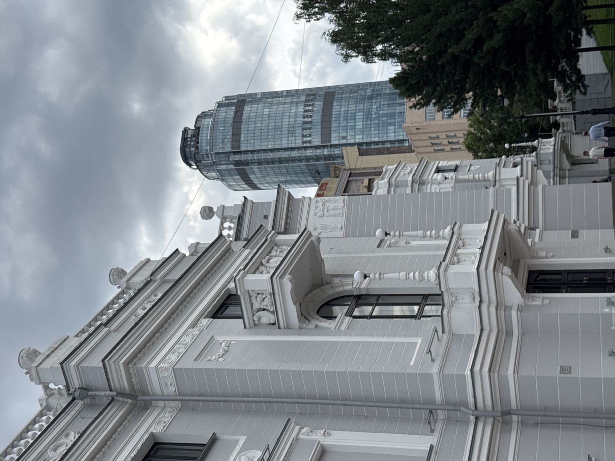 The image depicts the facade of a white, ornate building with classical architectural elements, such as columns, balconies, and decorative carvings, juxtaposed against a modern, cylindrical glass skyscraper in the background. The sky is overcast with heavy, gray clouds. The white building has dark windows, a dark entrance door, and decorative railings. In front of the building, two men are walking away from the camera. The modern skyscraper, which appears to be Vysotsky Business Center, is tall and has a distinctive curved shape with blue-tinted glass windows. Trees are visible to the right of the modern skyscraper. The overall impression is a blend of historical and contemporary urban architecture under a somber sky in Yekaterinburg, Russia.