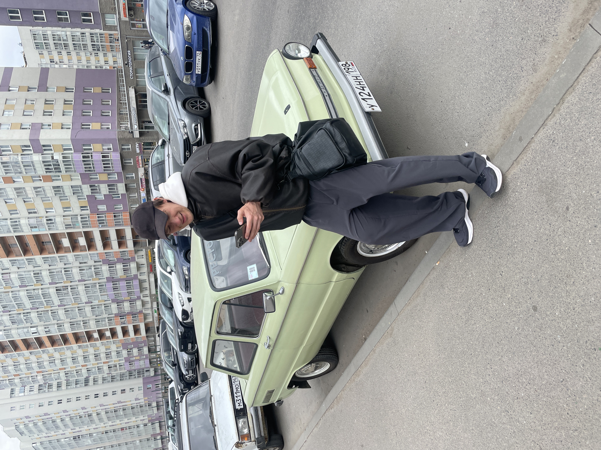 A medium shot depicts a man leaning on a light green ZAZ-968 Zaporozhets car parked on a city street in an urban area in Unknown, Russia. The man, facing the camera, wears a black jacket over a white hoodie, dark grey pants, a black cap, and black and white sneakers. He holds a smartphone in his left hand, seemingly taking a photo. A black messenger bag is slung over his shoulder.
The ZAZ-968 has the license plate У 124 НН 198. Several other parked vehicles are visible in the background, including a blue BMW with license plate У 011 УК 72 and a white Lada. The background includes a multi-story apartment building with a mixed color palette, predominantly white, grey, and purple. Some storefronts with signs are also visible. The pavement is dry and gray.
