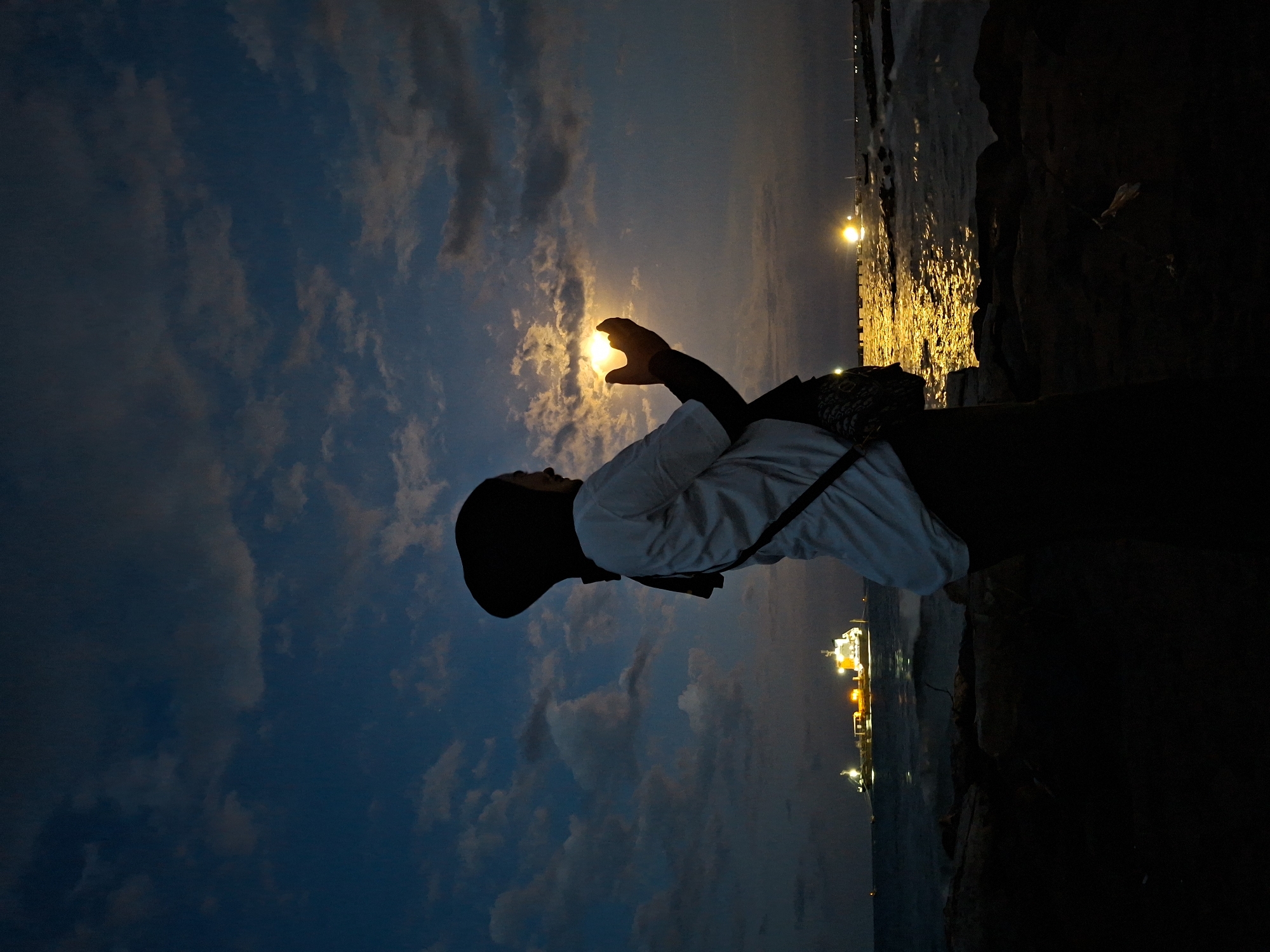 Here's a forensic description of the image:
A low-angle, landscape-oriented shot taken at night depicts a person silhouetted against a cloudy sky. The person is wearing a head covering (hijab), a light-colored long-sleeved shirt, and dark pants. They are standing on what appears to be a rocky shoreline, with dark rocks visible on the right edge of the frame. The person is holding their hand up towards the moon which appears as a bright orb nestled within the cupped hand. Beyond the person, a body of water reflects lights, possibly from ships or shoreline structures. The sky transitions from a dark blue at the top to a lighter hue near the horizon. The image was taken in Lhokseumawe, Indonesia.