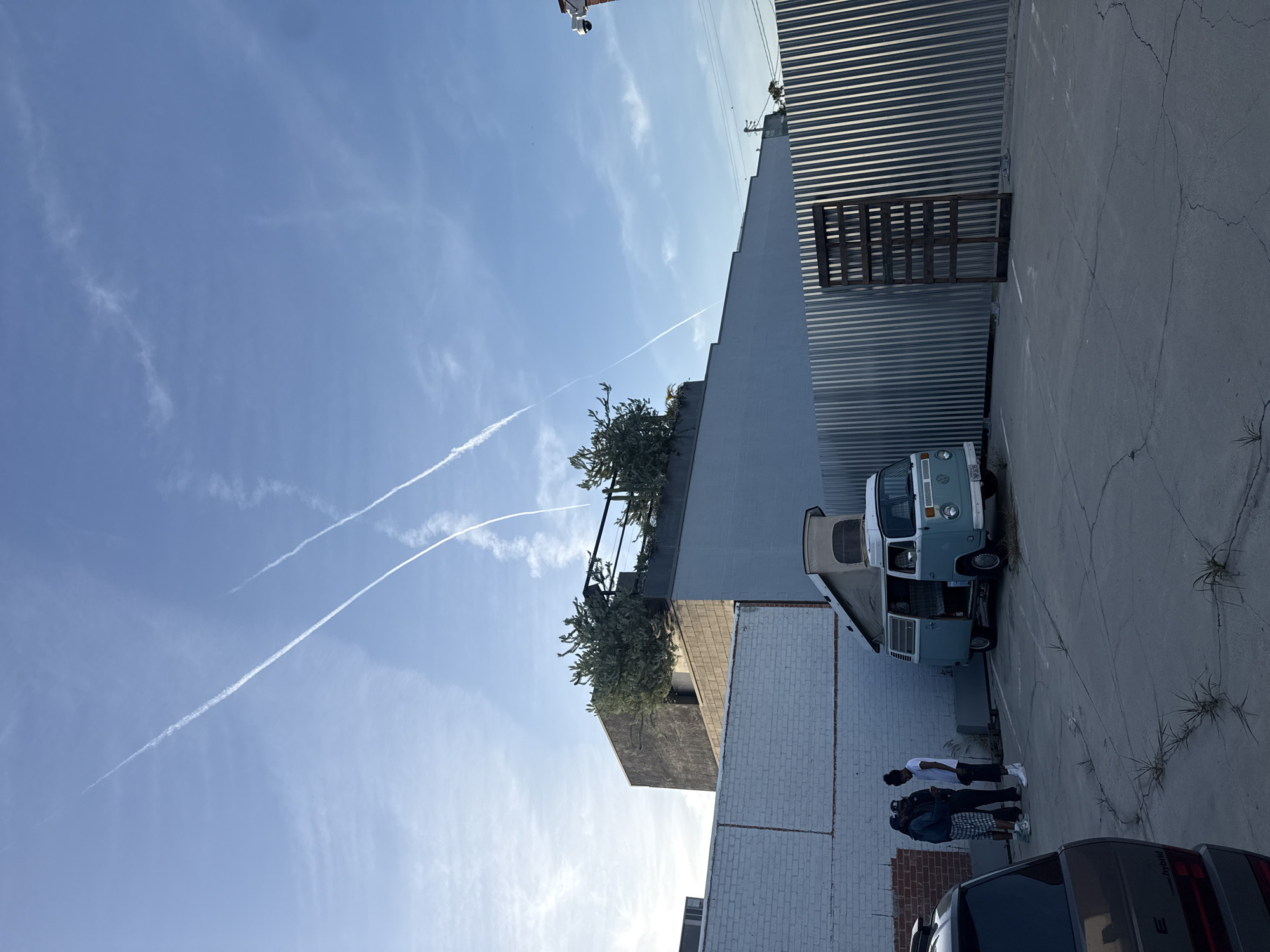 The image shows a partly cloudy sky over a parking lot in what appears to be an urban area in Culver City, California. Three visible contrails bisect the clear blue sky. A blue and white vintage Volkswagen van is parked adjacent to a corrugated metal wall and a white brick building. Three people stand near the van. The rear of a dark-colored minivan is visible at the bottom of the image. A tall gray building is adjacent to the VW. Plants grow on the roof of the tall building.