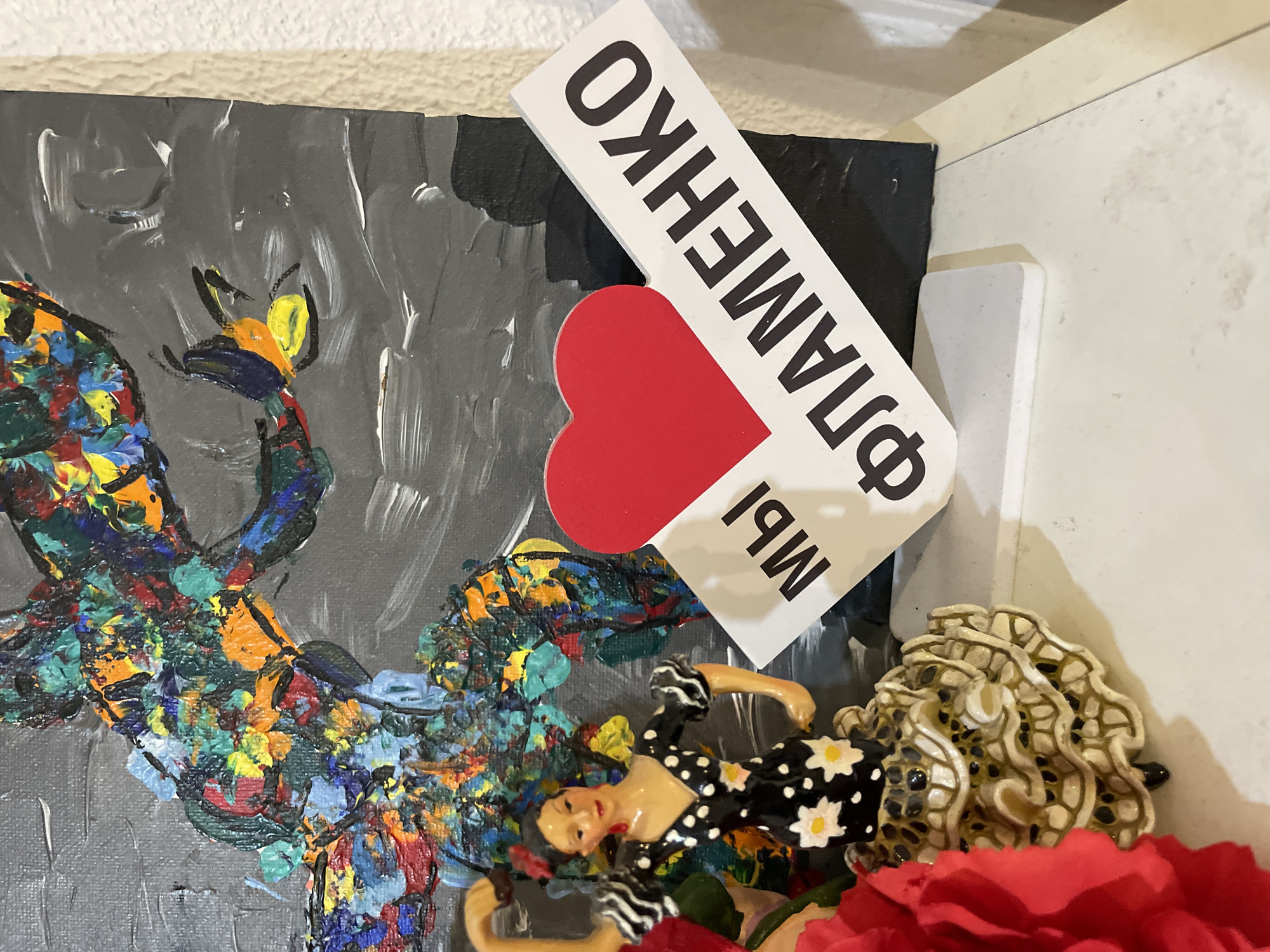This media file captures a close-up of a decorative arrangement, likely on a shelf or display in Sochi, Russia. The scene features a mixed-media display of art and figurines.
A central element is an abstract painting with a heavily textured, impasto style. Its background is a dark gray with visible white brushstrokes, providing a stark contrast to the vibrant, thick dabs of blues, greens, reds, yellows, and oranges that form an organic, branch-like or abstract shape.
Overlapping the painting is a white, rectangular sign tilted diagonally. It features a red heart sticker in the middle. The sign displays black Cyrillic text, which, when read correctly (it appears inverted in the image), spells out "МЫ ФИЛАМЕНКО" (MY FILAMENKO), translating to "WE ARE FILAMENKO" or "WE, FILAMENKO."
Below the sign and to the right, a small figurine of a woman lies on her back. She has pale skin, dark hair styled in an updo, and wears a black dress adorned with white polka dots and white daisy-like flowers. Her pose suggests a relaxed recline. Adjacent to her is a cream-colored, intricately textured object, possibly a decorative shell or ceramic piece. In the bottom right corner, a vibrant red artificial flower, resembling a rose or carnation, adds a pop of color to the arrangement. The overall impression is a personal and artistic display. No specific activity is occurring; it is a static collection of objects.