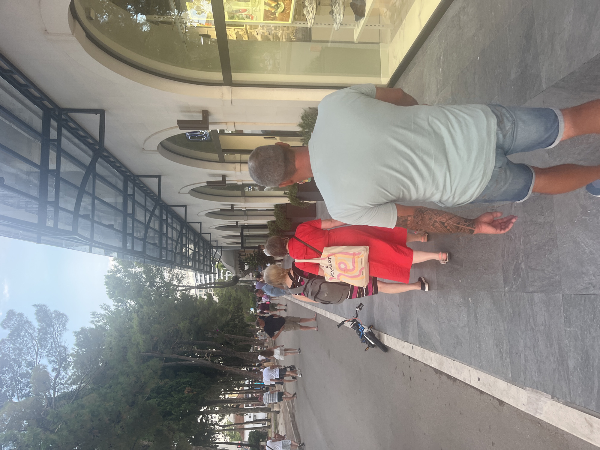 In an outdoor urban setting, a man with visible arm tattoos walks away from the camera on a grey tiled sidewalk next to a street with several people strolling. He wears a light blue t-shirt and denim shorts. To his left, a woman in a red dress and carrying a tote bag walks in the same direction, followed by another woman with a backpack. Further down the street, a bicycle lies on its side on the pavement near the curb. On the right side of the image, a building features large arched windows showcasing merchandise, possibly shoes, and a dark awning extends over the windows. Tall trees line the left side of the street. The sky is visible in the upper portion of the image, with some clouds.