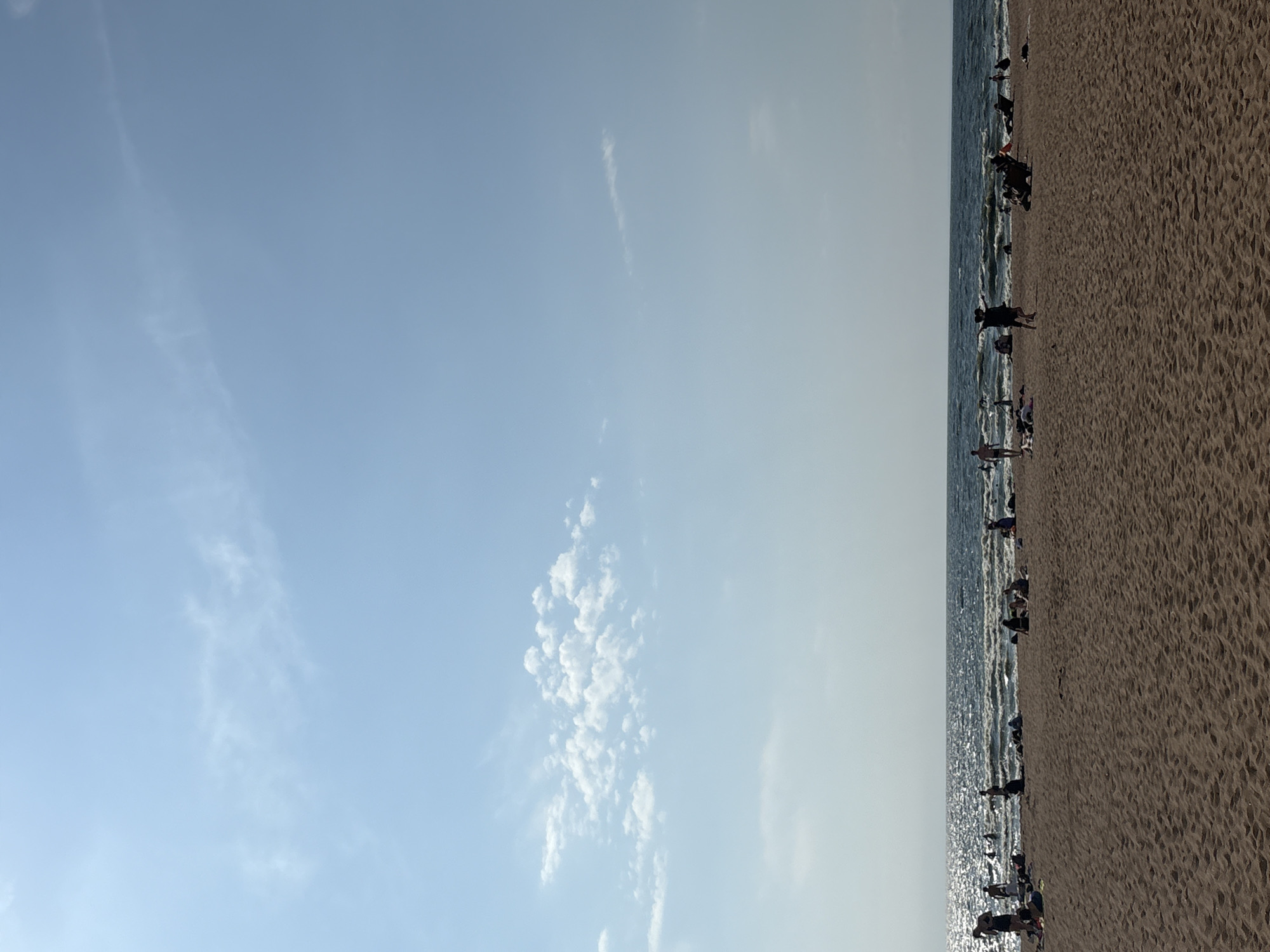 The image depicts a sunny day at a sandy beach in Solnechnoye, Russia. The foreground is dominated by the textured sand of the beach, with tire tracks visible. In the middle ground, numerous people are scattered across the sand and in the shallow water. Some are lying down, others are standing or walking, and a few are playing in the waves of the Baltic Sea. The sea itself is a deep blue, with white-capped waves reflecting the bright sunlight. The horizon line is clear, with a light haze softening the distant view. The upper portion of the image is filled with a pale blue sky, featuring scattered white clouds, one large cluster of cumulus clouds and a few wispy cirrus clouds. The overall atmosphere is one of leisure and recreation.