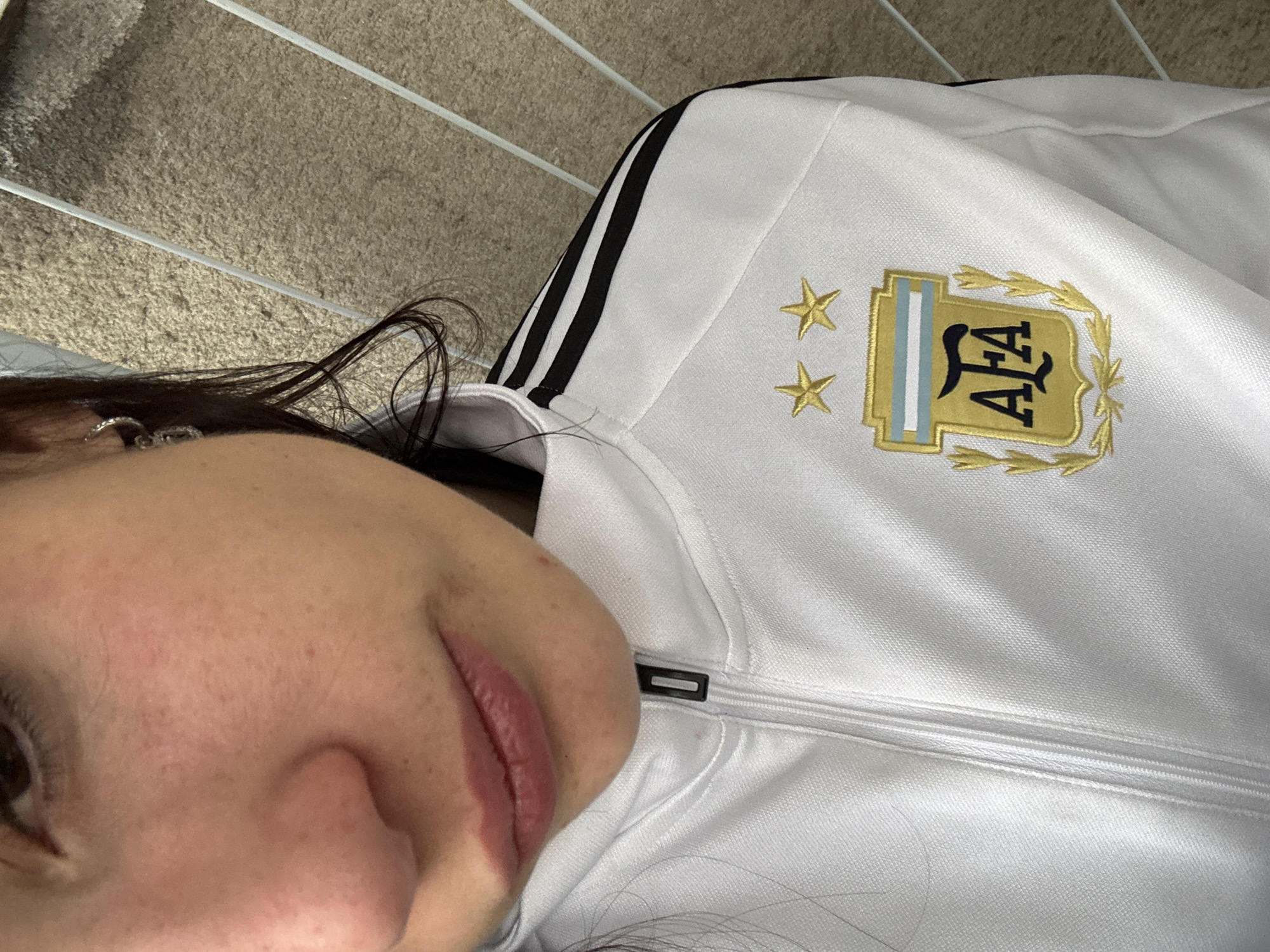 A close-up, tilted shot focuses on a woman's face, partially visible, wearing a white zip-up jacket. The jacket features black stripes on the shoulders and the Argentina national football team logo, indicating it is a sports garment. The background reveals a carpeted area. The Argentina football team's logo is situated on the upper chest area of the jacket. The context suggests Argentina because of the soccer jacket logo, but the photo does not contain any location information. Therefore, I can’t provide any specific location within Buenos Aires.