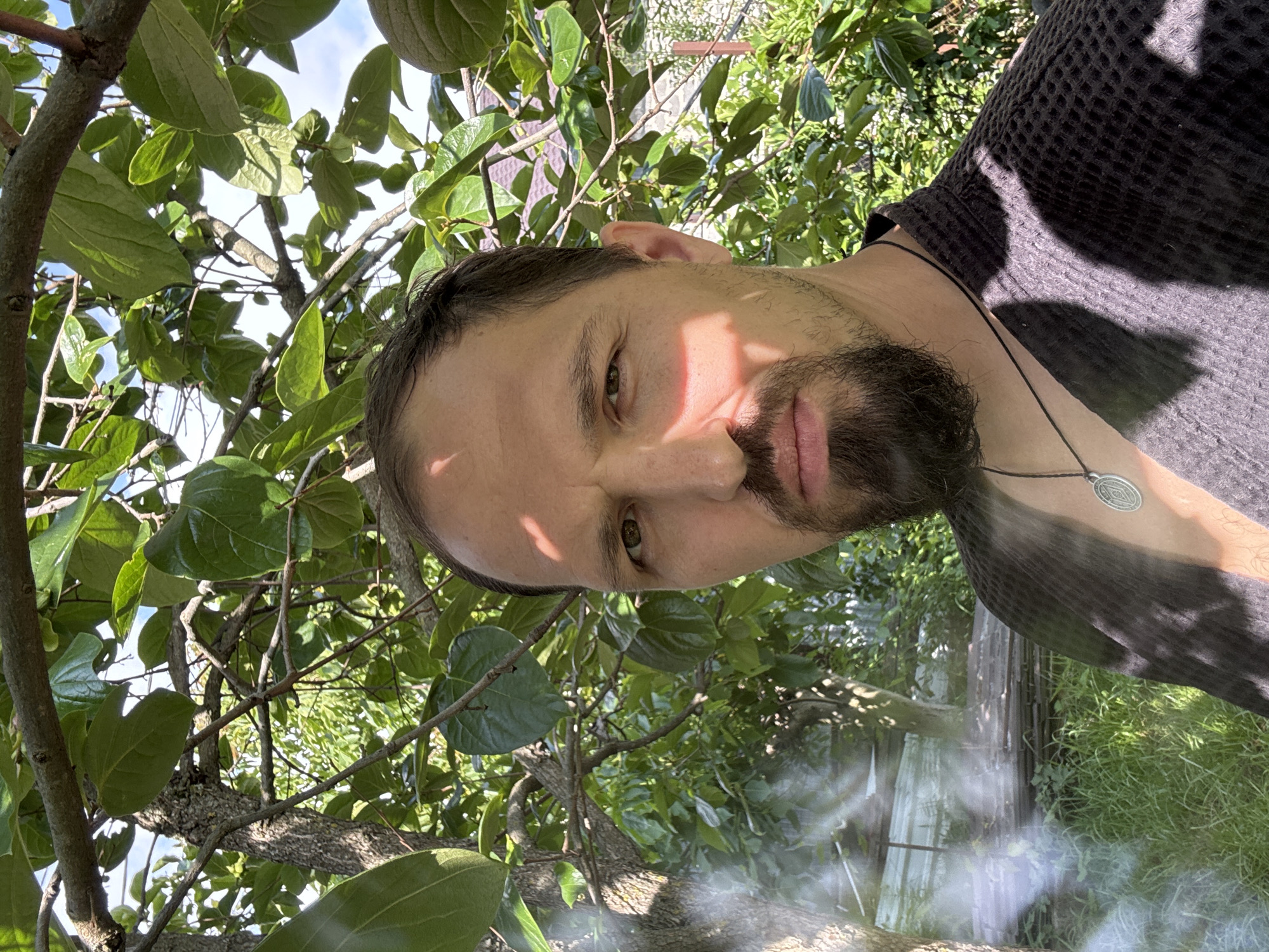 An extreme close-up selfie shows a man with a goatee and a serious expression, partially obscured by tree branches and leaves. He is wearing a dark, textured robe or jacket with a necklace featuring a circular pendant. Sunlight casts dappled shadows across his face and clothing. The background suggests an outdoor setting, possibly a garden or yard, with lush green foliage and what appears to be a building with a tiled roof in the distance. The image's lighting and composition create a somewhat dramatic and intense mood. The location is not specified in the image itself, but it's stated to be in Sochi, Russia.