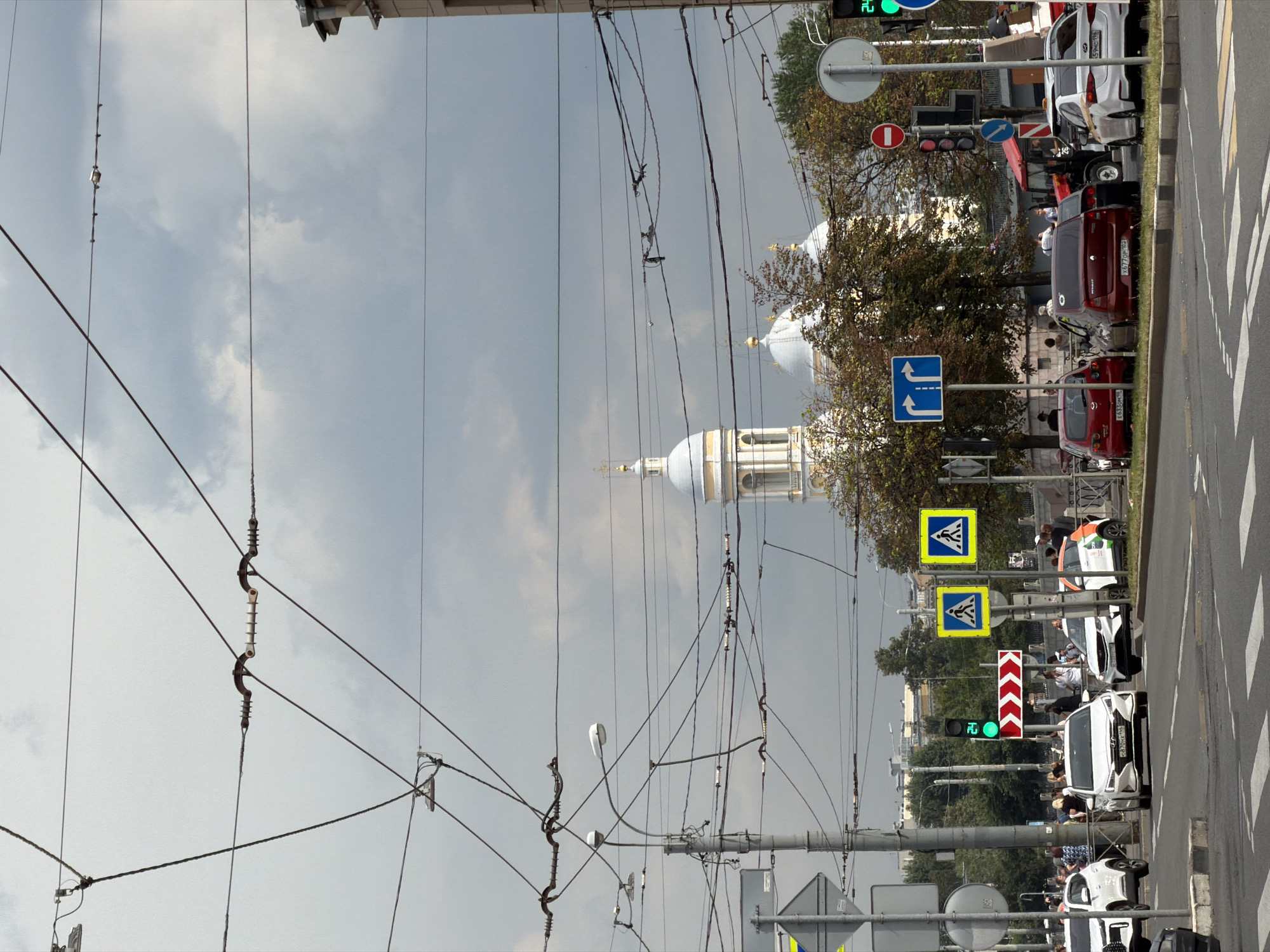 This image depicts a street scene in Saint Petersburg, Russia, with a prominent view of the Smolny Cathedral and its bell tower in the background. The foreground is dominated by a complex network of overhead wires, likely for public transportation such as trams or trolleybuses, crisscrossing the frame against a cloudy sky. Several cars are visible on the road, including white sedans, a red hatchback, and a white car with an orange and green decal. Pedestrians are present on the sidewalks and near a crosswalk. Traffic signals, road signs, and streetlights are also visible, indicating an active urban environment. The architecture of the Smolny Cathedral, with its distinctive blue and white domes and golden spires, serves as a recognizable landmark within the city. The overall atmosphere is that of a typical day in a bustling Russian city, with a focus on the interplay between urban infrastructure and architectural heritage.