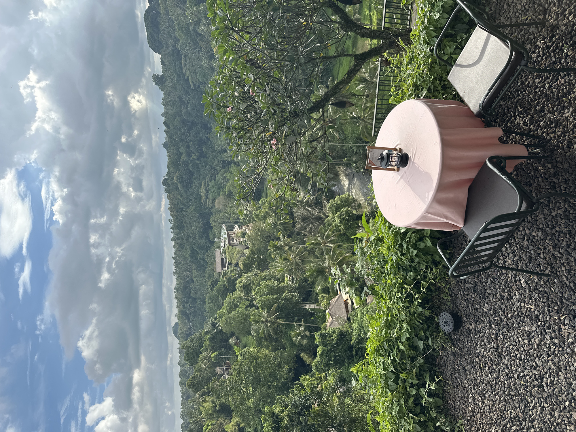 The image presents a lush landscape scene in the Ubud District, Indonesia. A table covered with a pink cloth sits on a gravel surface, accompanied by two chairs. The table is decorated with a lantern. The vantage point provides a view overlooking a dense, tropical forest, with occasional structures nestled amongst the trees. In the background, a cloudy sky looms. The overall scene suggests a peaceful, scenic outdoor setting at a dining or rest area.