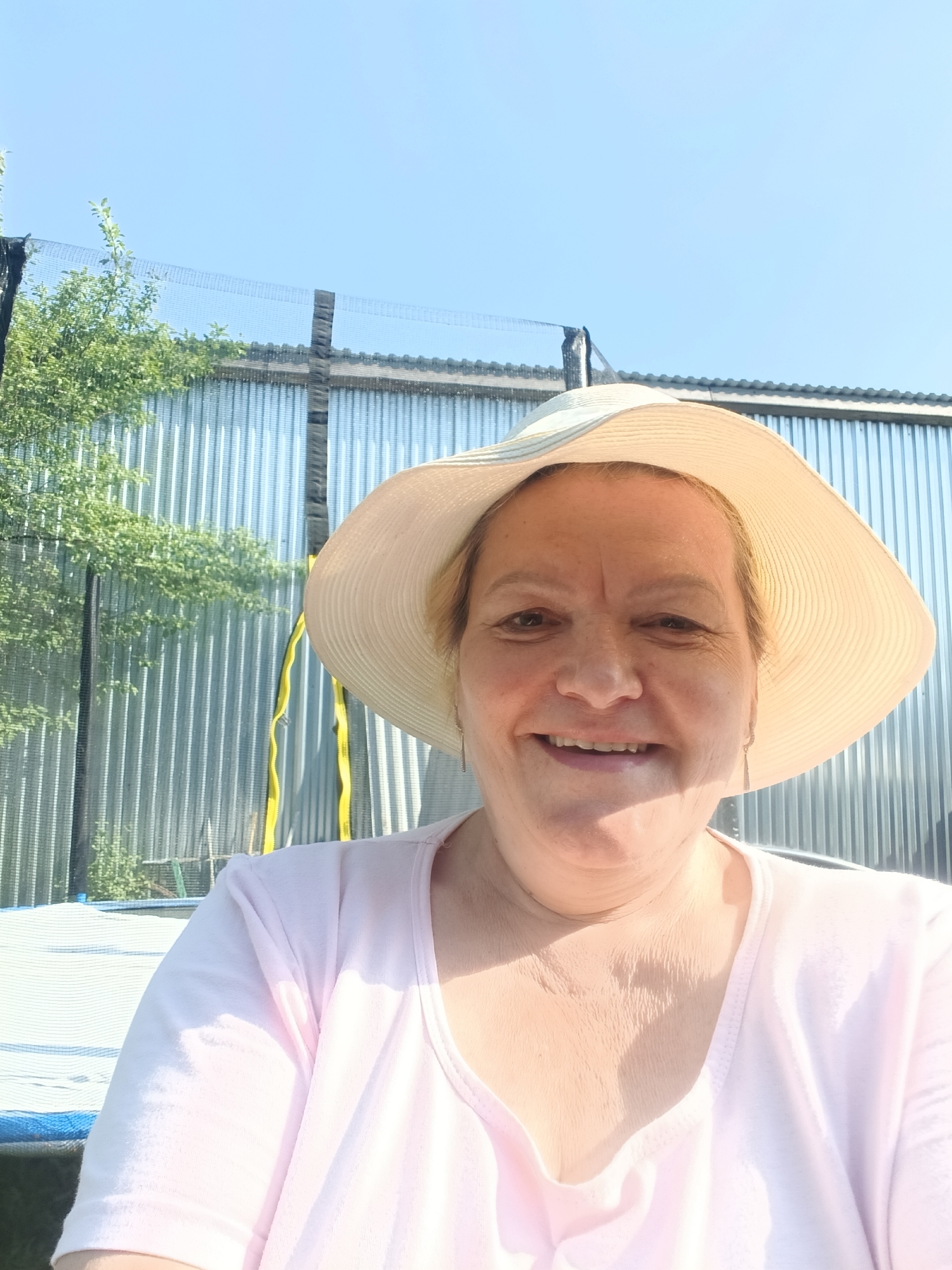 A middle-aged woman with fair skin and blonde hair is smiling at the camera while wearing a wide-brimmed hat and a light pink t-shirt. Behind her is a corrugated metal fence with a trampoline partially visible on the left. A leafy tree is also visible to the left of the fence. The sky is clear and blue. The image appears to be taken outdoors on a sunny day. There is no specific information indicating the location is in the city of Красный Маяк, Russia.