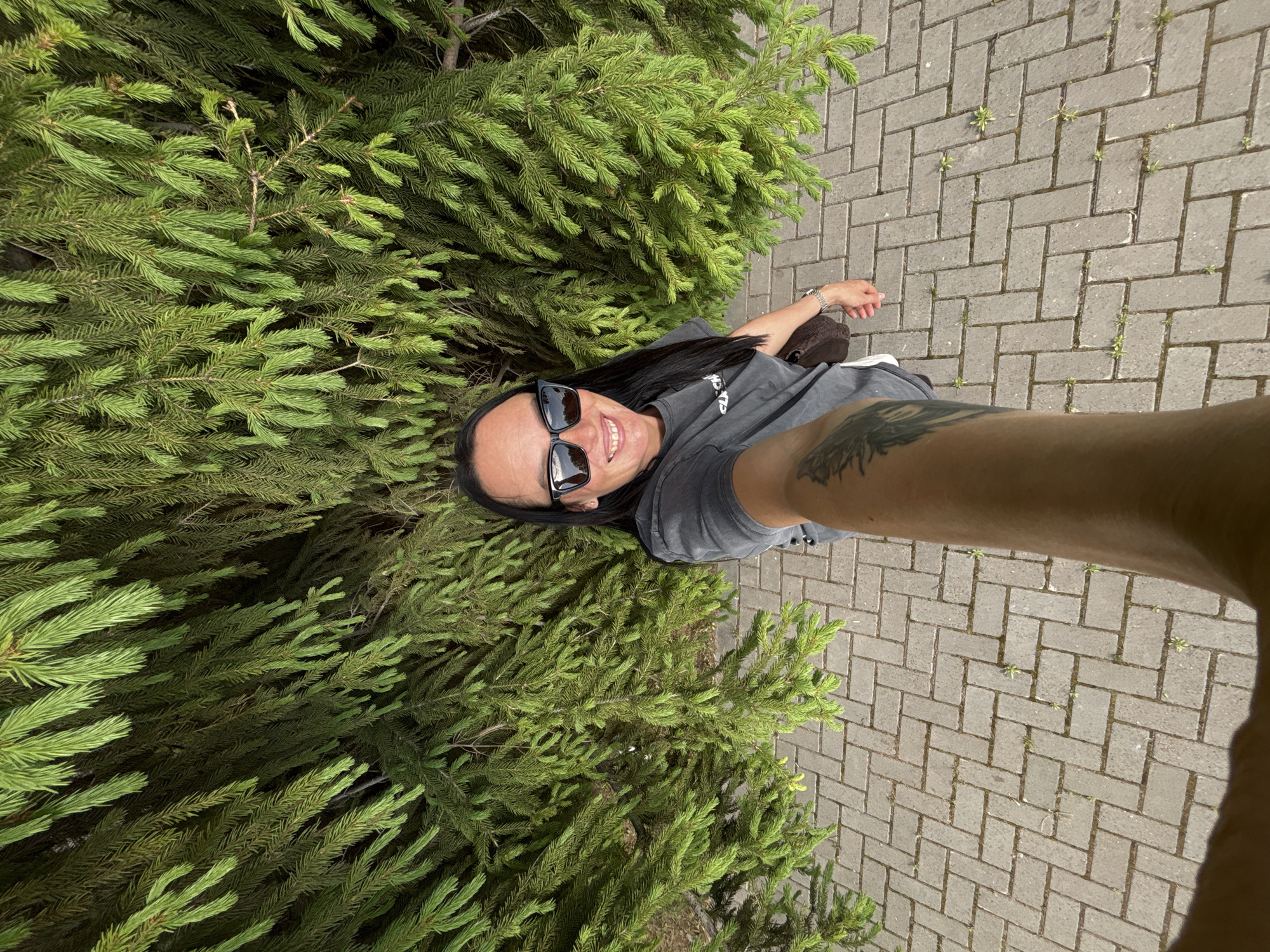 A woman is taking a selfie outside in Izhevsk, Russia. She is wearing sunglasses and a t-shirt. Her arm with a tattoo is visible in the foreground. A fir tree is on the left and a brick sidewalk is on the right.