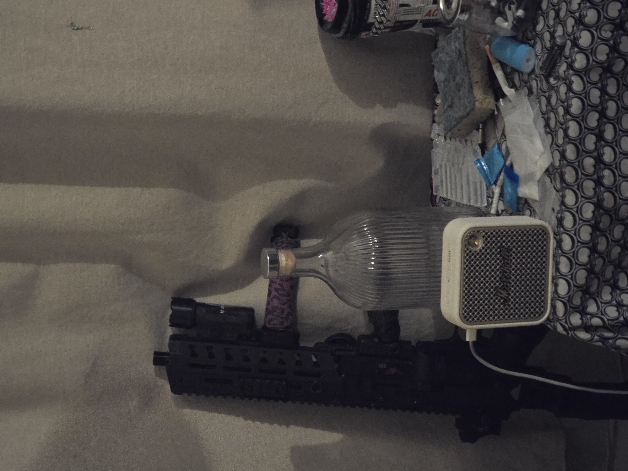 The image shows a cluttered scene with various objects. A black assault rifle with a flashlight is visible at the bottom. Above it is a glass bottle with a cork. Next to the bottle is a small, off-white speaker. Other items in the image include cleaning supplies such as sponges and cleaning cloths. There are also small, blue packets, and a can of something is present at the top. A patterned fabric is also visible. There is no evidence to place the scene in Paris, France.
