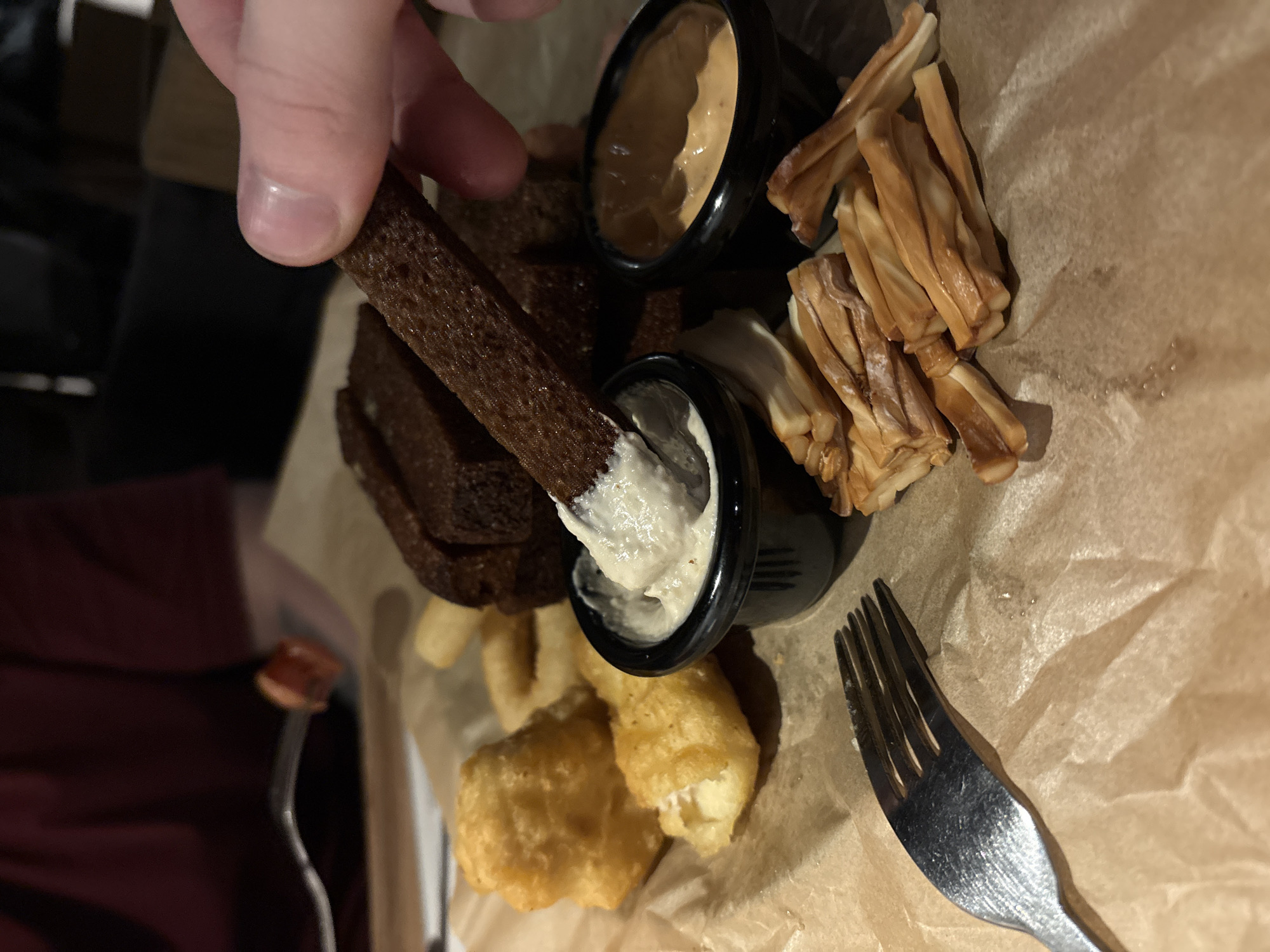 A close-up shot depicts a hand dipping a dark brown piece of bread into a small black container of white sauce. The bread is one of many pieces beside the container. A second black container holds a tan sauce. Cheese sticks rest beside the second container. Several pieces of fried food are shown at the bottom of the frame near a fork. All items are on wrinkled brown paper. The location is the city of Chaikovsky, Russia.