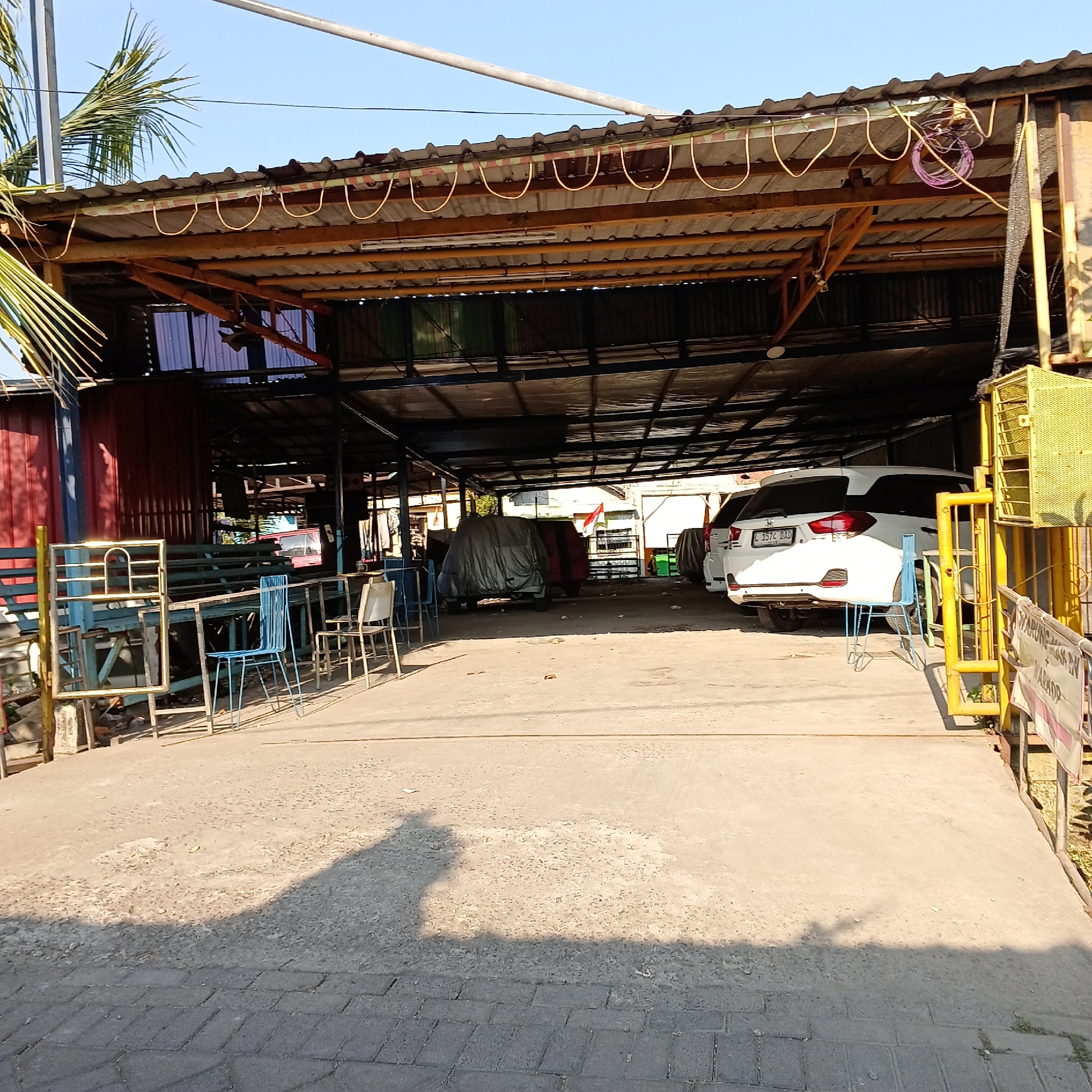 The image depicts an outdoor covered area, likely a car wash or workshop, in Wedoro, Indonesia. A white SUV with license plate "L 1574 DAD" is parked on the concrete floor. In the background, other vehicles are visible, some covered with tarps. Various metal structures, tables, and chairs are scattered around the area. A yellow metal gate and a sign are positioned on the right side of the frame. The scene is illuminated by sunlight, casting shadows on the ground. The overall environment suggests a functional, open-air commercial space in a tropical setting.
