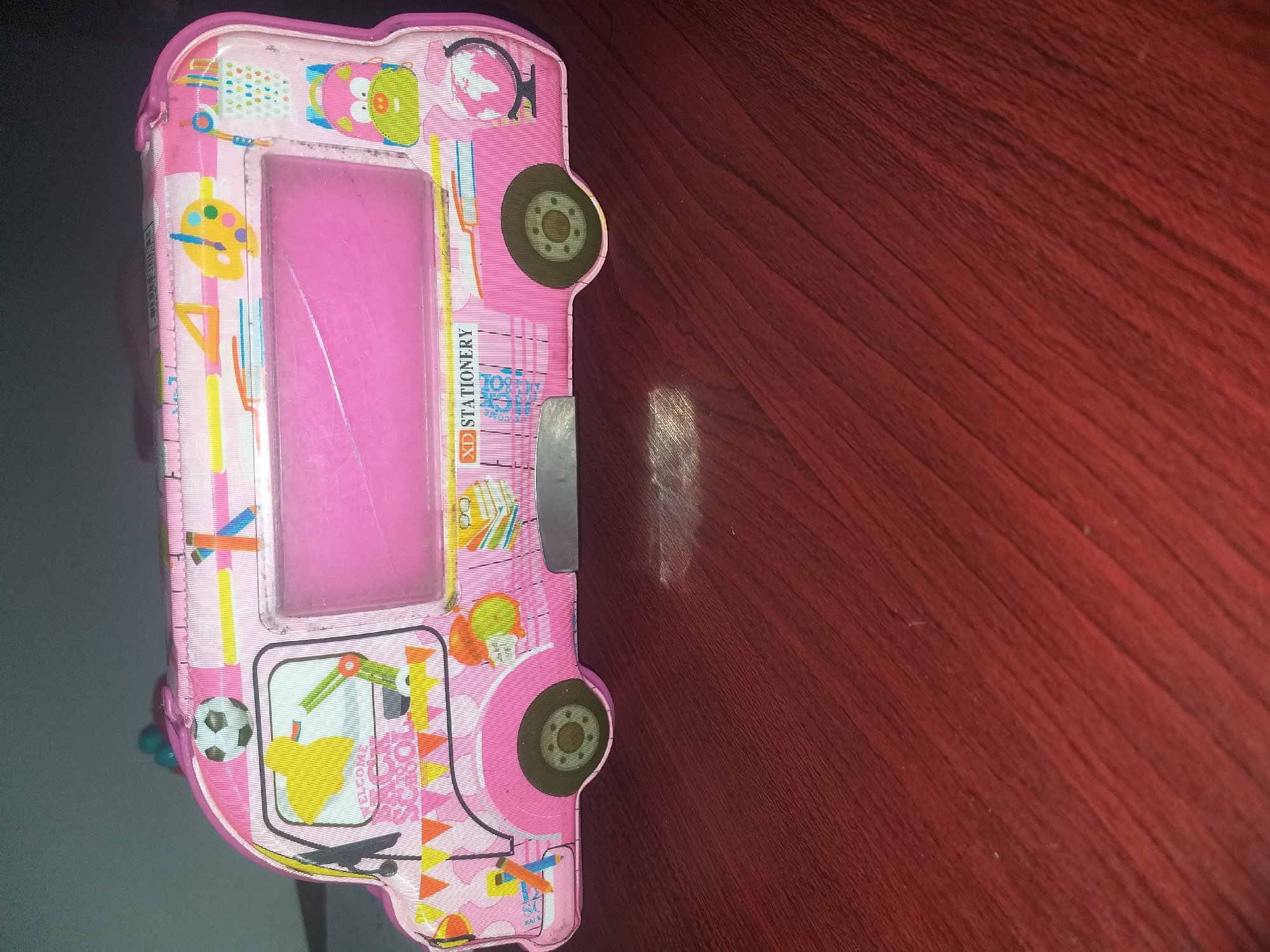 The image shows a pink pencil case designed to look like a school bus, resting on a dark red wooden surface. The pencil case features various colorful illustrations of school-related items, such as a soccer ball, a palette with paints, scissors, a globe, and books. It also has a clear plastic window on its side. The lighting in the image is dim, and there is a blurred reflection of light on the wooden surface in front of the pencil case. There are no people or animals visible in the image. The specific location of Air Jamban, Indonesia is not discernible from the image.