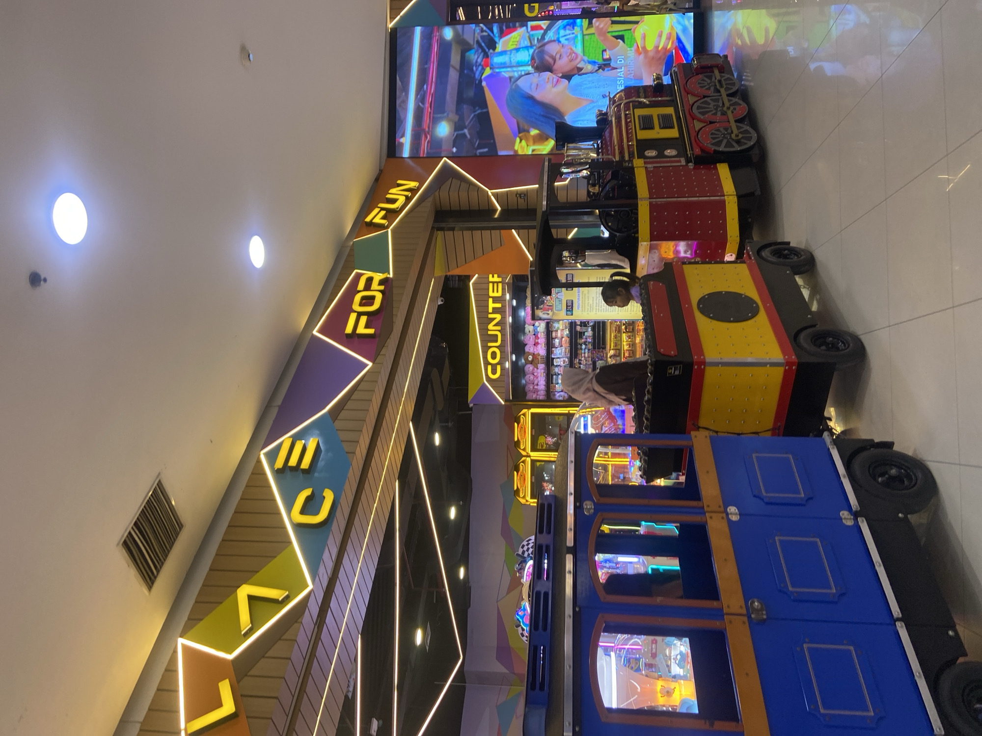 This image depicts a brightly lit interior of an amusement center, likely a game arcade or family entertainment center, in Sidoarjo, Indonesia. In the foreground, two small, colorful toy trains are stationary. The train on the left is primarily blue with yellow accents and wooden window frames. The train on the right is red with yellow and black accents, featuring visible wheels and detailed train car design.
Several people are present but not actively engaged in riding the trains. A woman wearing a hijab is standing near the red train, appearing to observe the surroundings. A young child is seated in the red train. In the background, a counter labeled "COUNTER" is visible, with shelves stocked with plush toys. Beyond that, various arcade games with glowing screens are illuminated, suggesting an active gaming area.
The environment is modern and well-maintained, with a clean tiled floor reflecting the ambient light. Overhead, decorative lighting fixtures and a large screen displaying colorful graphics contribute to the vibrant atmosphere. The signage above the trains includes the words "FOR FUN" in large, stylized yellow lettering. The overall scene conveys a sense of entertainment and leisure, typical of a family-friendly attraction.