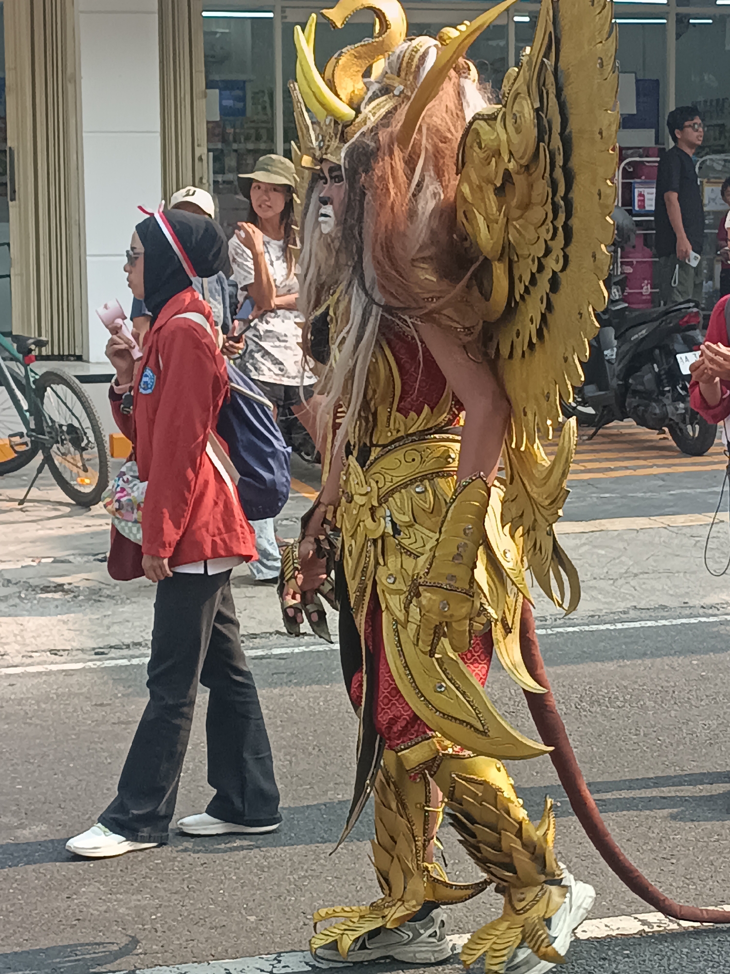 In Sleman, Indonesia, a street scene unfolds with a person in elaborate golden mythological creature costume walking on a paved road. The costume features intricate golden armor, large feathered wings, a long mane, and a tail. To the left, a young woman in a red jacket and black pants is walking, holding a pink object. Behind her, other individuals are visible, including a woman in a hat and a man wearing glasses and a black t-shirt. A bicycle is parked on the left side of the road, and a motorcycle is present in the background. The environment appears to be an urban street with buildings lining the side.