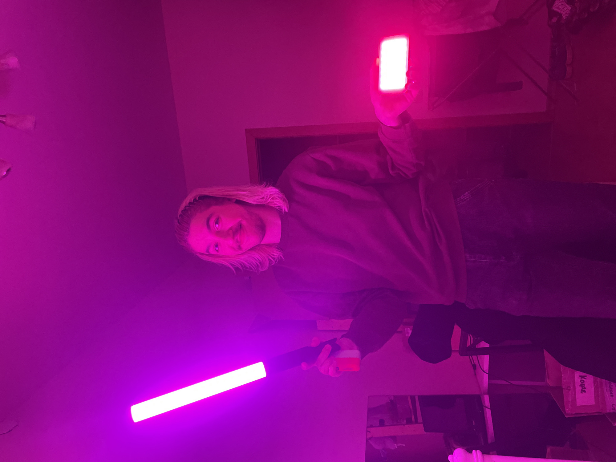 A person with long blond hair, wearing a dark sweatshirt and jeans, stands in a room illuminated by purple light. They hold a light-up tube in their left hand and a bright white phone or similar device in their right hand. The room contains furniture, including a desk, a chair, and a box labeled "Kopie". The location is not Moscow, Russia.
