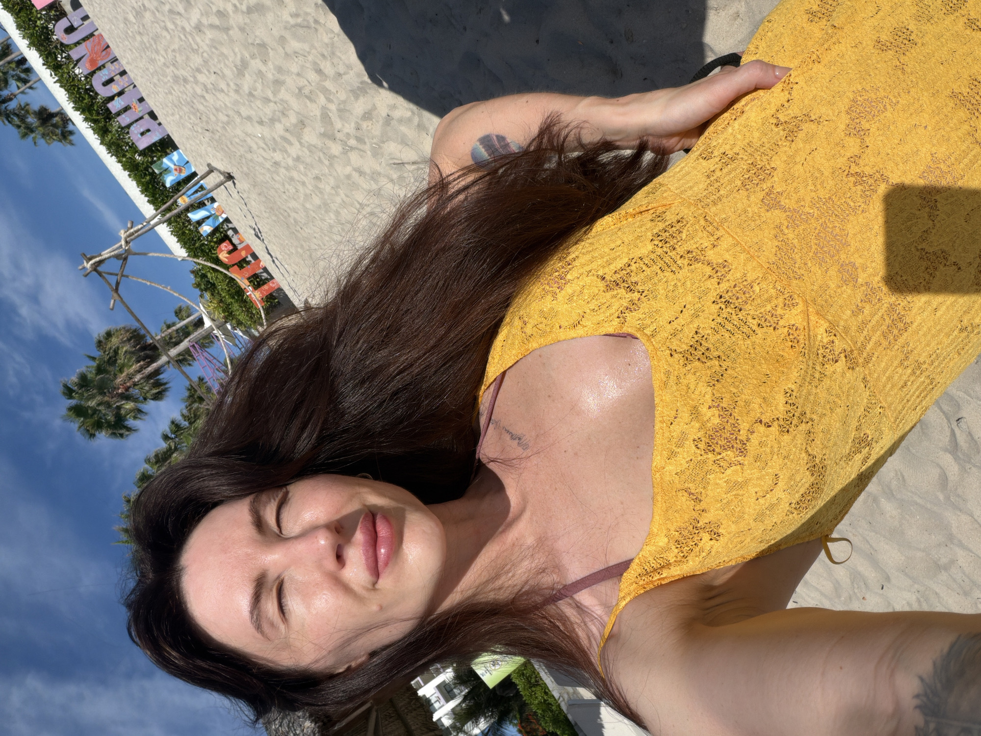 A young woman with long dark hair and closed eyes is lying on a sandy beach. She is wearing a yellow lace dress over a pink bikini top. Her skin is glistening with sweat, suggesting a warm, sunny day. In the background, palm trees line the shore, and a large sign with colorful letters spelling out "TIEN THANH PHONG" is visible. The sign appears to be a landmark or decoration for the beach area, which is located in Dong Ninh Hoa Ward, Vietnam. The overall scene conveys a sense of relaxation and enjoyment of a tropical vacation.