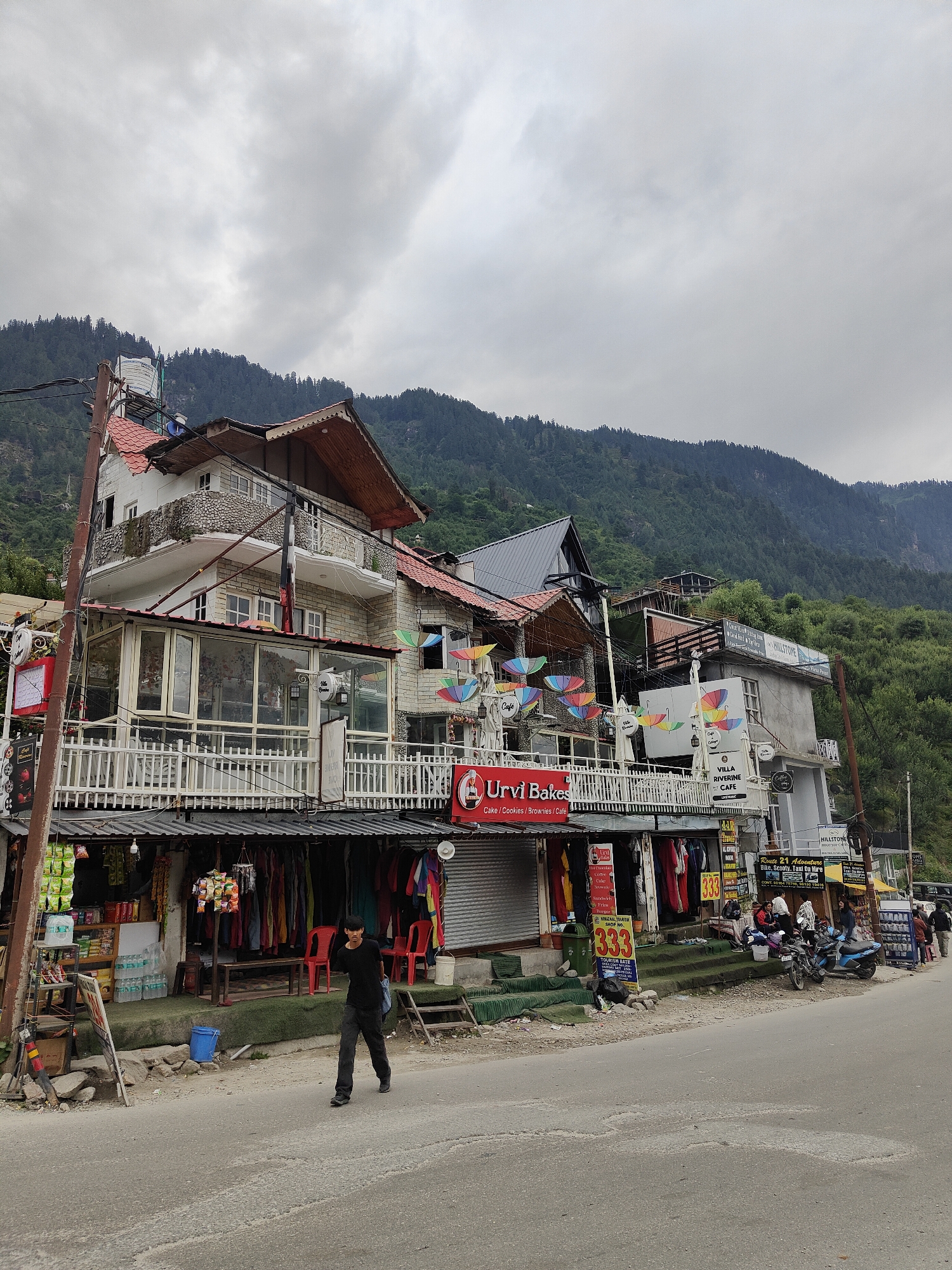 In Vashisht, India, on a cloudy day, a person walks down a street in front of a multi-story building with shops on the ground floor. The building has a white facade with stone accents and red-tiled roofs, and features balconies with white railings. Colorful umbrellas are displayed on the balconies of the upper floors. The ground floor shops are open, displaying clothing and food items. A man in a black t-shirt and dark pants walks on the asphalt road, carrying a blue bag over his shoulder. To the right of the shops, several motorcycles and scooters are parked, and a few people are gathered. Behind the buildings, a densely forested hillside rises steeply, with the sky a uniform gray.