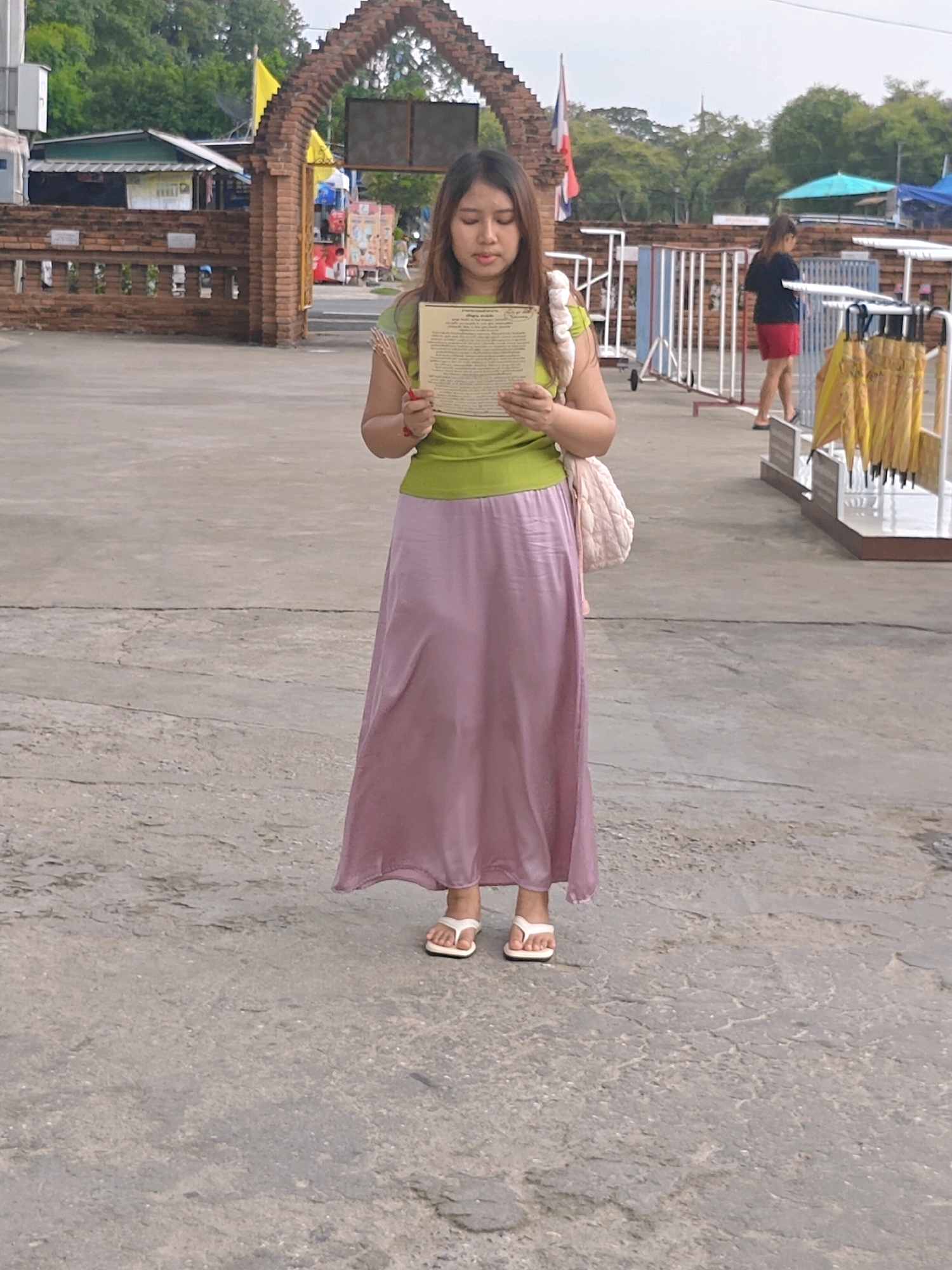 A young woman stands in a paved outdoor area, holding a piece of paper and a bundle of incense sticks. She is wearing a lime green t-shirt and a long, flowing lavender skirt. Her hair is dark and shoulder-length. A light pink quilted bag is slung over her left shoulder. She is looking down at the paper in her hands.
In the background, there is a brick archway adorned with a yellow flag. Further behind the archway, there are several buildings, some with awnings. A person wearing a black top and red shorts is visible on the right side of the image, walking away from the camera. A rack of yellow umbrellas is also present to the right. The ground is made of concrete, showing some cracks and wear. The overall environment suggests an outdoor public space, possibly near a temple or market in Phra Nakhon Si Ayutthaya City Municipality, Thailand. The lighting is overcast, indicating a cloudy day.