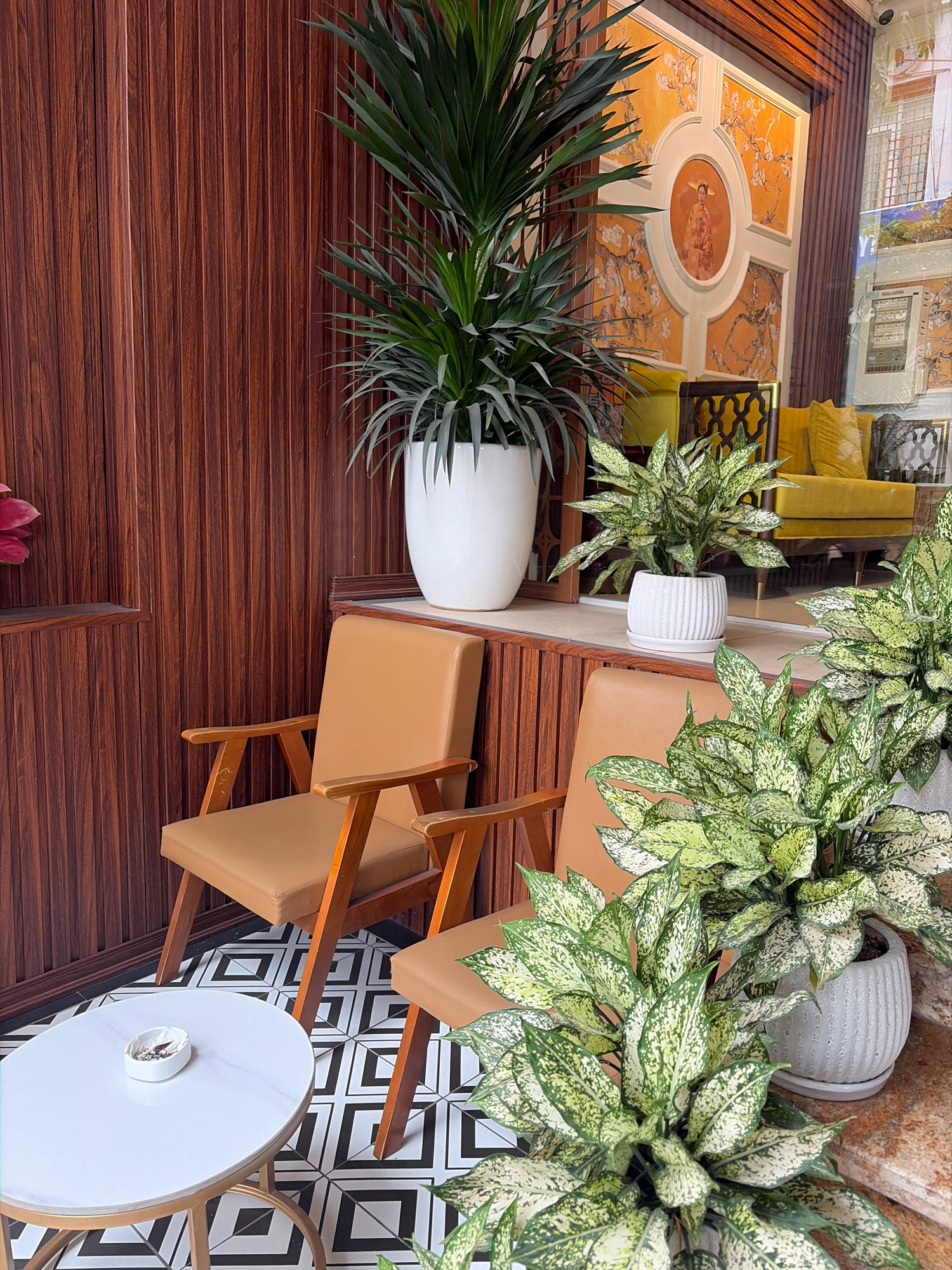 The image captures an indoor seating area, likely a cafe or lounge, with a strong emphasis on plants and decorative elements. Two brown leather chairs with wooden frames are positioned around a small, oval white table with a marble-like surface, resting on a gold base. The table has an ashtray with cigarette butts on it. The floor features a black and white geometric tile pattern.
Behind the chairs, a tall, leafy green plant in a large white pot stands against a dark brown wooden paneled wall. To the right of this, on a wooden shelf, another plant in a white ribbed pot is visible, with a plush yellow sofa and chairs in the background, framed by a large window or reflective surface.
The back wall is adorned with an ornate mural featuring a central circular depiction of a person in traditional attire, surrounded by a yellow patterned motif of branches and flowers. The overall aesthetic suggests a stylish and contemporary setting, possibly in a commercial establishment in Hanoi, Vietnam, given the architectural and decorative style often found in such spaces. There are no people visible in the image.