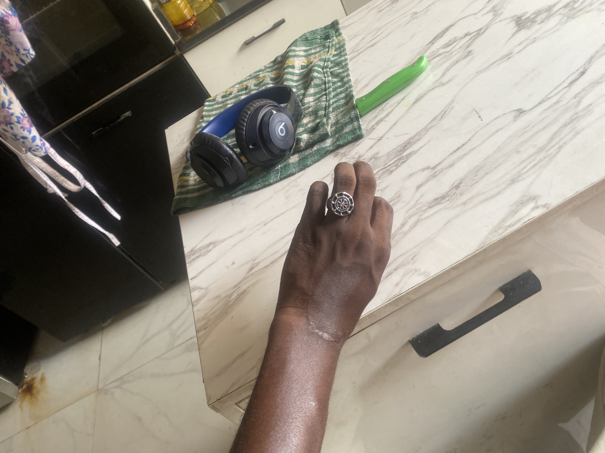 The image captures a domestic scene set in Lagos, Nigeria. A dark-skinned arm and hand, adorned with a silver ring on the ring finger, prominently occupy the foreground. The arm shows a scar on the wrist. On a marble-patterned countertop, a pair of blue and black Beats headphones rest on a striped green and white towel, alongside a green-handled knife. Behind the countertop, dark kitchen cabinets are visible, with a glimpse of a white-tiled floor below. The overall scene suggests an indoor setting within a home or apartment.