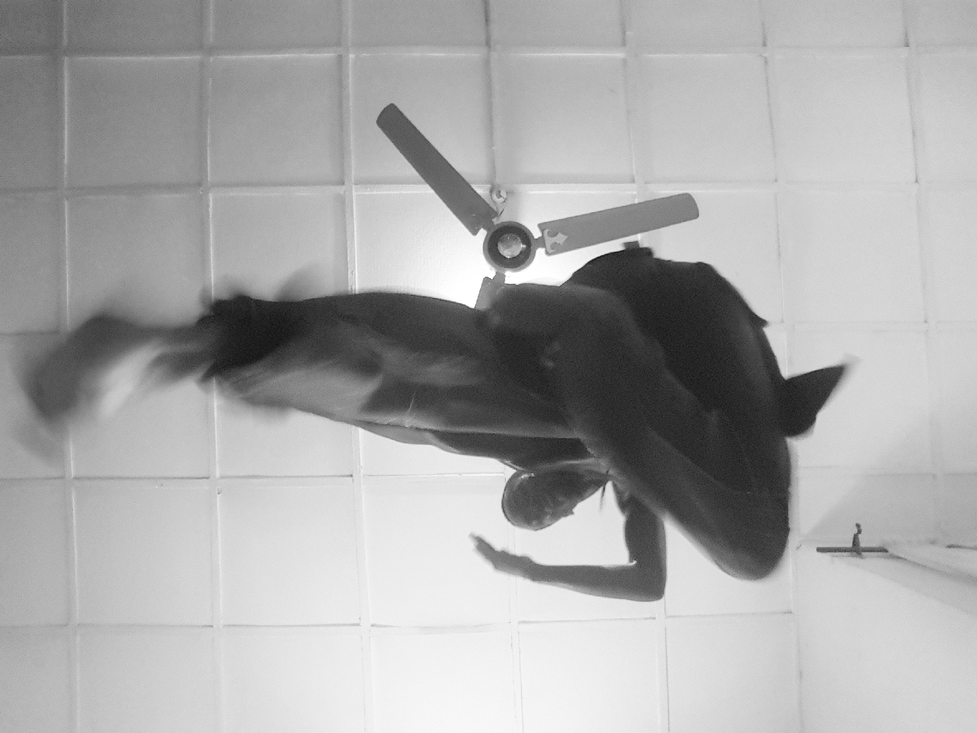 This black and white image, captured in Narayi, Nigeria, depicts a single individual in mid-action, photographed from a low-angle perspective looking directly upwards. The individual is centrally positioned, appearing inverted relative to a standard upright orientation, with legs extending towards the top of the frame and the head and upper torso towards the bottom. Significant motion blur is evident across the entire figure, particularly in the limbs, indicating rapid movement. The individual is wearing dark clothing. The background consists of a light-colored, uniformly tiled ceiling with clearly visible grid lines. A three-bladed ceiling fan is mounted on the ceiling on the right side of the frame, with its blades appearing relatively stationary compared to the blurred figure. A dark, vertical structural element, possibly a door frame or wall edge, is partially visible at the bottom left, featuring a small, dark horizontal fixture.