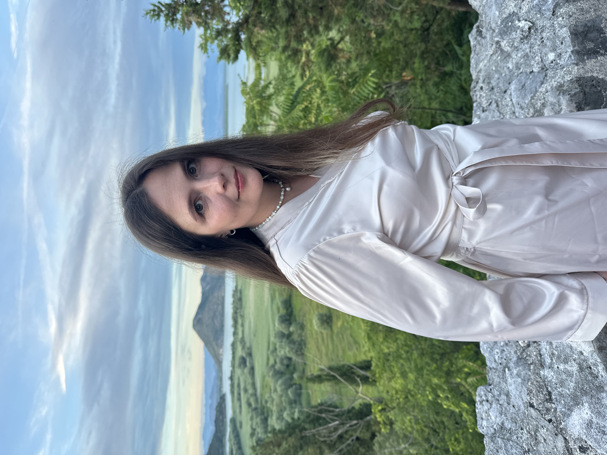 A young woman with long, brown hair stands in front of a scenic view of Skadar Lake, near Virpazar, Montenegro. She is wearing a light-colored, satin robe with a pearl necklace. The landscape behind her features rolling green hills, a tranquil lake, and distant mountains under a cloudy sky. The woman is positioned on a stone wall, looking towards the camera with a slight smile. The scene is captured during twilight, indicated by the soft lighting and atmospheric clouds.