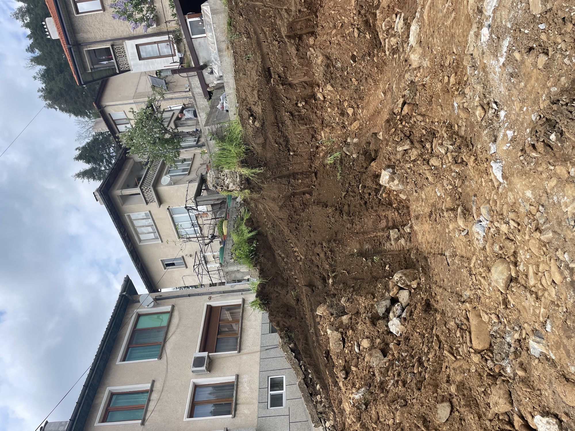 The image depicts an excavation site located adjacent to residential buildings, likely in Zlatograd, Bulgaria. The foreground is dominated by a large, irregular trench dug into reddish-brown soil containing numerous rocks and debris. Embedded wooden supports, possibly for shoring, are visible along the trench's edge. Adjacent to the excavation, several multi-story buildings are situated, their facades showing signs of age and diverse architectural styles, including balconies, windows with varying frames, and external air conditioning units. The sky is overcast. There is no evidence of people in the frame.