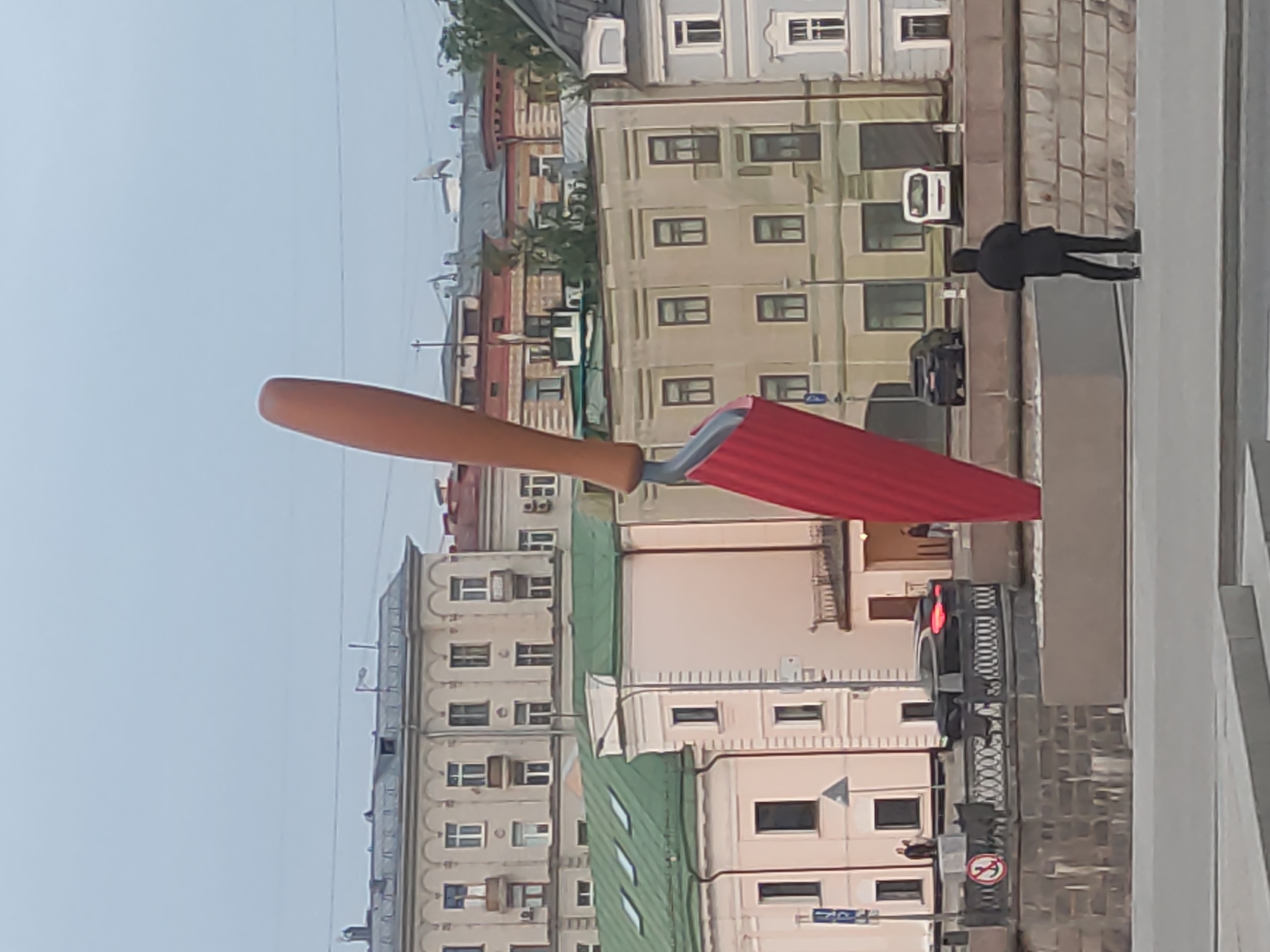 A vertical, eye-level shot depicts a large, likely public art installation of a red and brown masonry trowel, set against a backdrop of Moscow city buildings. The trowel is positioned upright on a gray stone ledge, with a pedestrian walking by on the sidewalk to the right. The city buildings in the background consist of multi-story, beige and gray structures with numerous windows and some greenery interspersed. Overhead power lines are visible against a gray sky.