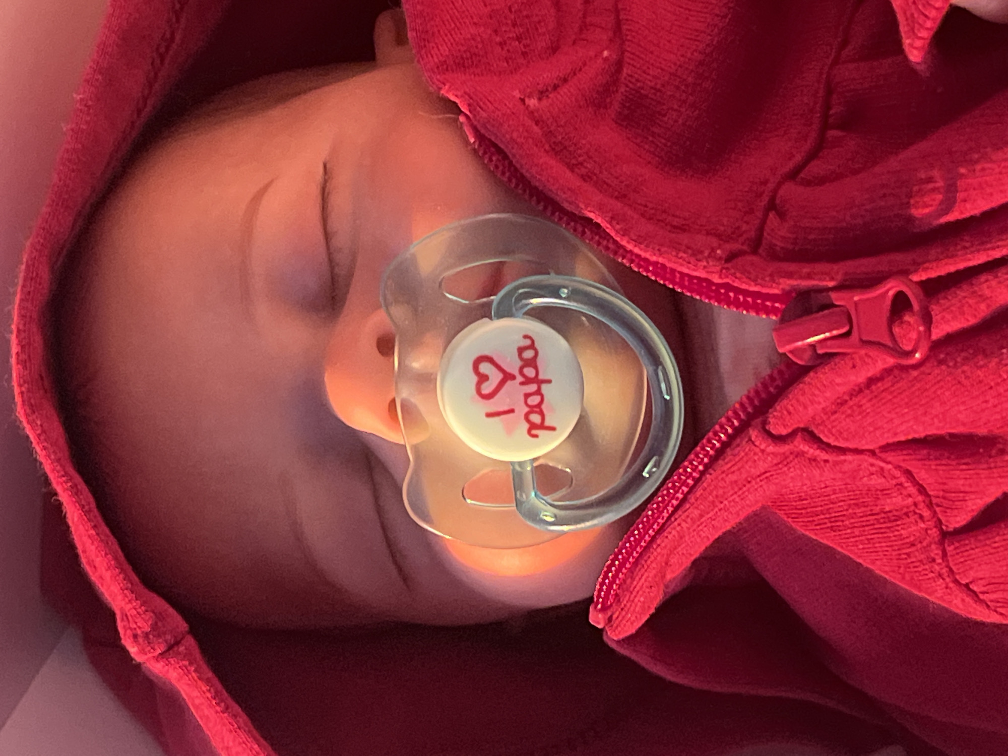 The media file shows a close-up, overhead shot of a sleeping baby wearing a red hooded garment. The baby has a pacifier in its mouth, which features a white button with a red heart and the text "I ❤ papa" written in red script. The baby is nestled within the red garment, suggesting a sense of warmth and security. The lighting appears soft and possibly artificial, creating a warm, intimate atmosphere. There are no discernible outdoor or environmental cues to indicate the time of day, weather, or specific location within Votkinsk, Russia. The overall emotion conveyed is peacefulness and contentment, due to the baby's closed eyes and relaxed posture. The focus is entirely on the baby and the pacifier, with the red garment providing a vibrant backdrop.