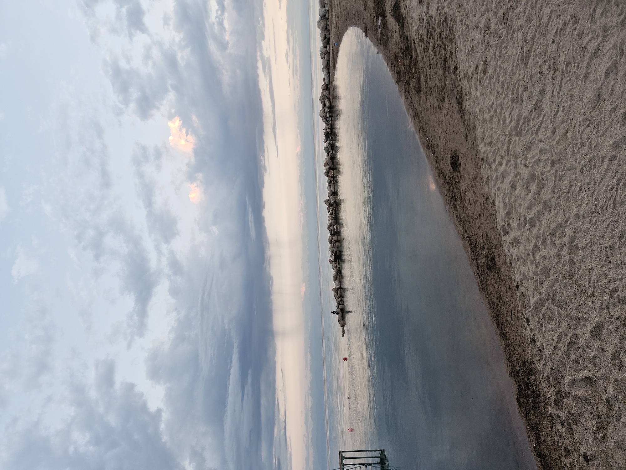 A wide-angle, eye-level shot captures a serene lake scene at dusk near Bulan-Segettu, Kyrgyzstan. The sky is a canvas of muted blues and grays, with soft clouds reflecting the fading light. A patch of light pink clouds glows near the horizon, hinting at the sun's descent. The lake's surface is calm, mirroring the sky and distant, snow-capped mountains that form a faint silhouette on the horizon.
In the foreground, a sandy beach curves along the water's edge. The sand is light brown and textured, with some scattered debris near the water line. To the left of the frame, a wooden structure, possibly a pier, extends into the water. Three buoys, one red and two pink, float on the lake's surface.
Further out on the lake, a breakwater made of large stones juts into the water, creating a sheltered cove. On top of the breakwater, a lone figure is seated, facing the water, suggesting a moment of quiet contemplation or fishing. The overall atmosphere is peaceful and tranquil, characteristic of a lakeside evening.