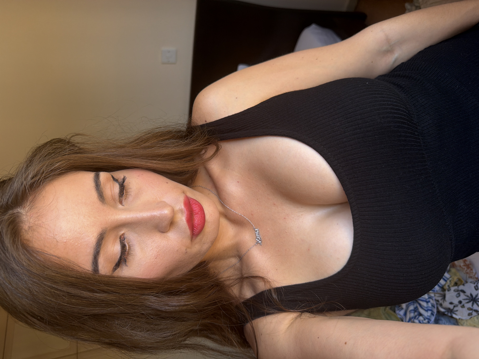 A close-up shot captures a woman with long, brown hair and fair skin, looking down with her eyes closed. She is wearing a black, ribbed tank top and a silver necklace with a pendant that reads "Mama." Her makeup includes bright red lipstick and winged eyeliner. The background is a plain, light-colored wall with a dark headboard and a white pillow visible on a bed. The scene appears to be indoors, possibly in a bedroom. There is no indication of the city of Tersefanou, Cyprus in the image.