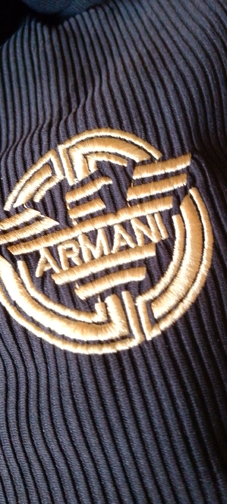 The image shows a close-up view of a dark blue ribbed fabric with a gold embroidered logo. The logo is circular with a design inside and the word "ARMANI" embroidered in the center. The fabric appears to be part of an article of clothing or accessory. The image does not provide any information about the city of Jalingo, Nigeria, or any specific location within it.