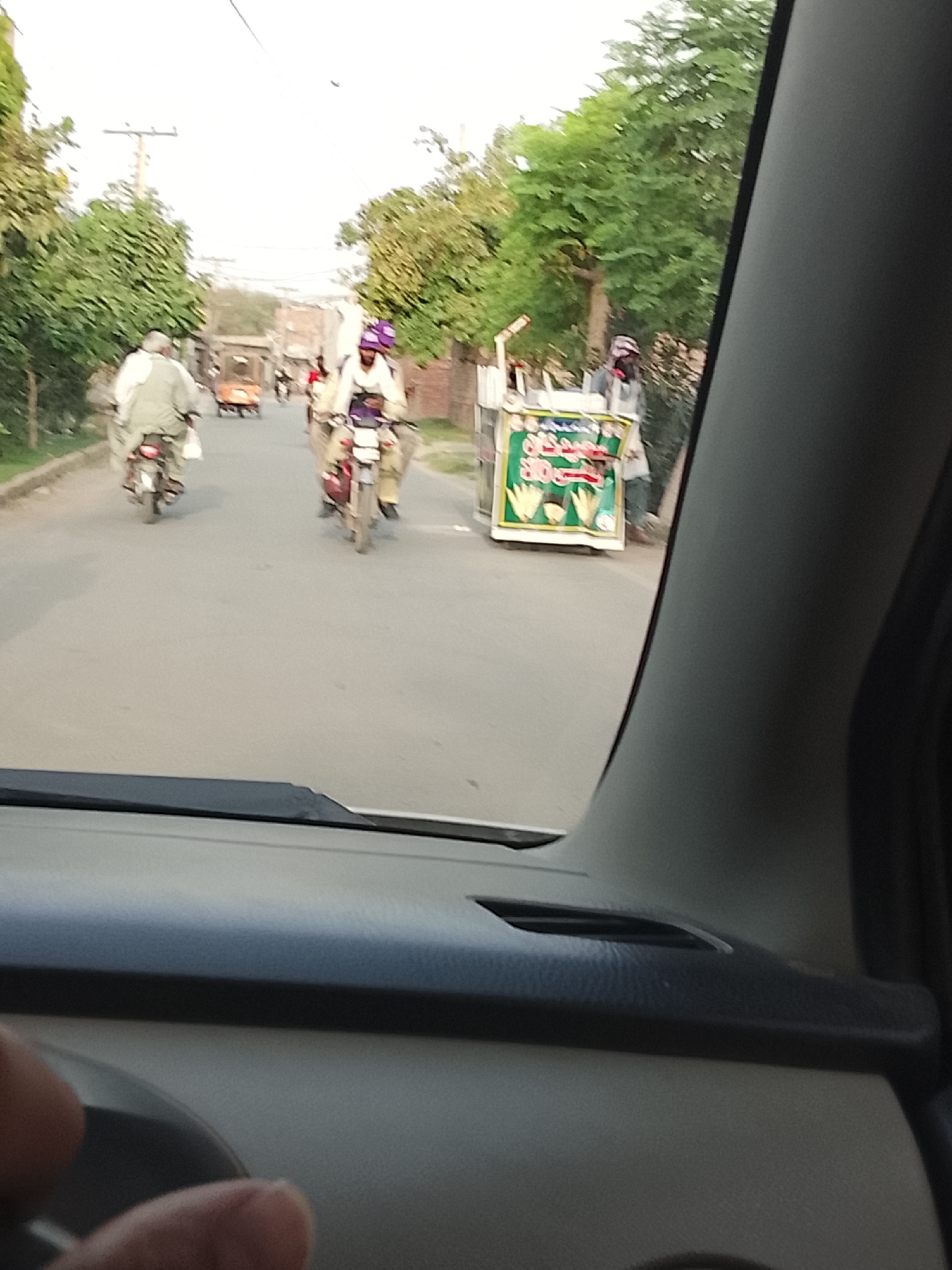 The image, taken from inside a vehicle, shows a street scene in what is possibly Nasheman Colony, Pakistan. In the middle ground, there are two motorcycles traveling in the same direction as the vehicle. The first motorcycle has a rider wearing white clothing, and the second has a rider wearing white and a purple helmet. Further ahead, a yellow vehicle is barely discernible. To the right, a food cart with green signage and images of corn is positioned on the side of the road. A man is standing near the cart. The environment consists of trees lining the street and buildings in the background. The sky is overcast. The foreground shows the dashboard and the interior of the vehicle from which the picture was taken.
