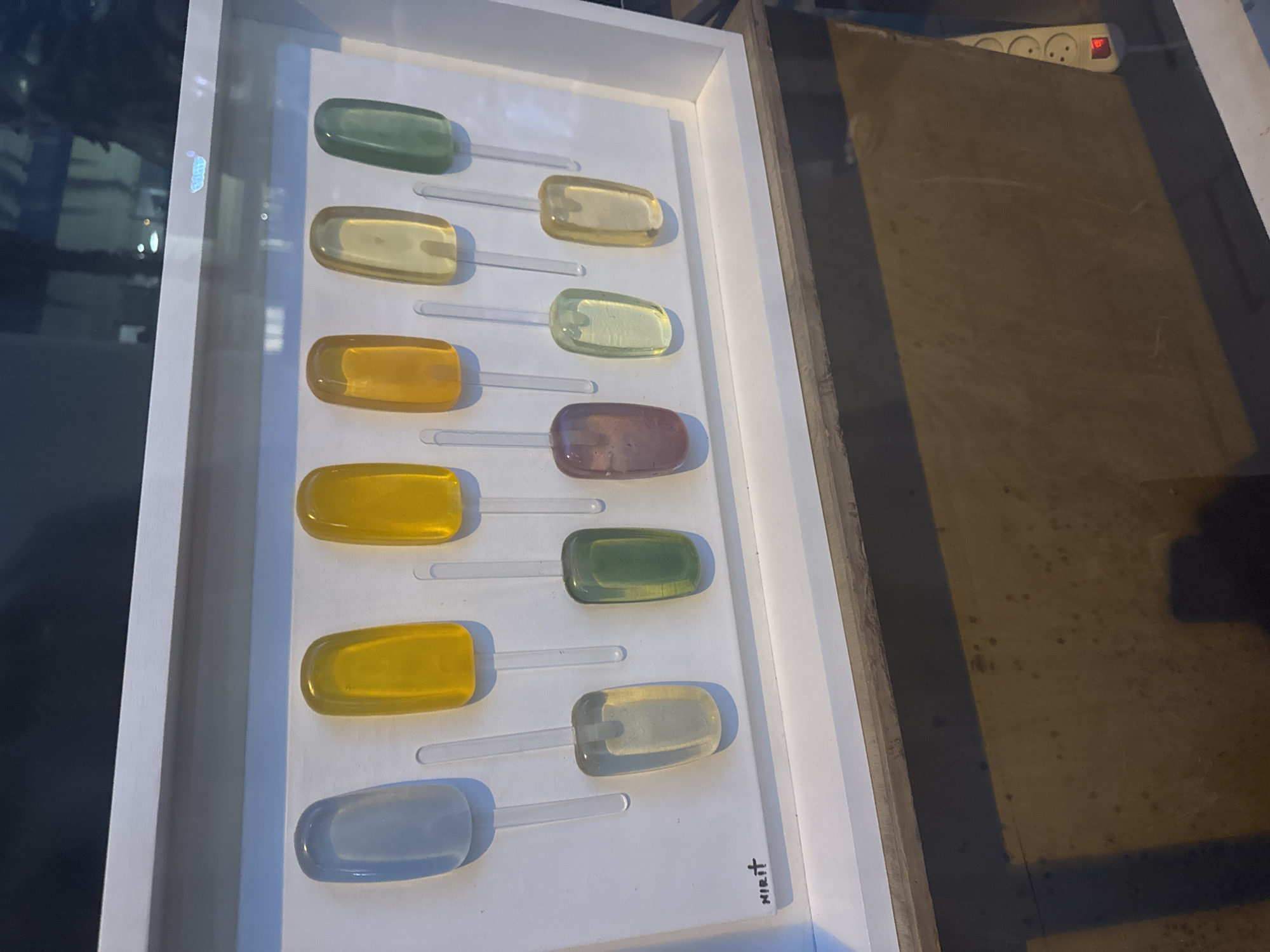 The image shows a framed art piece displaying a series of colorful, oblong shapes attached to clear sticks, resembling lollipops or candies. The "candies" are arranged in two columns, with varying colors including green, yellow, amber, purple, and blue. The background of the framed art is white.
The art piece is labeled "Mirit" in the lower right corner. The art is located in Tel-Aviv, Israel.
The image is taken indoors, with a glimpse of a power strip on a wooden shelf in the background. The photo appears to be taken at an angle, offering a view of both the artwork and the surroundings.