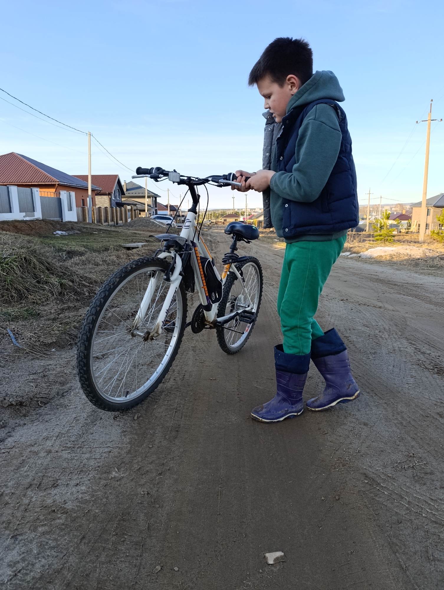 A young boy stands next to a bicycle on a dirt road in Chaikovsky, Russia. The boy is wearing a dark blue vest over a green hooded sweatshirt, green sweatpants, and dark blue rubber boots. He is looking down at the handlebars of the white and orange bicycle. The road is muddy with visible tire tracks. In the background are houses with red tile roofs and utility poles against a light blue sky.