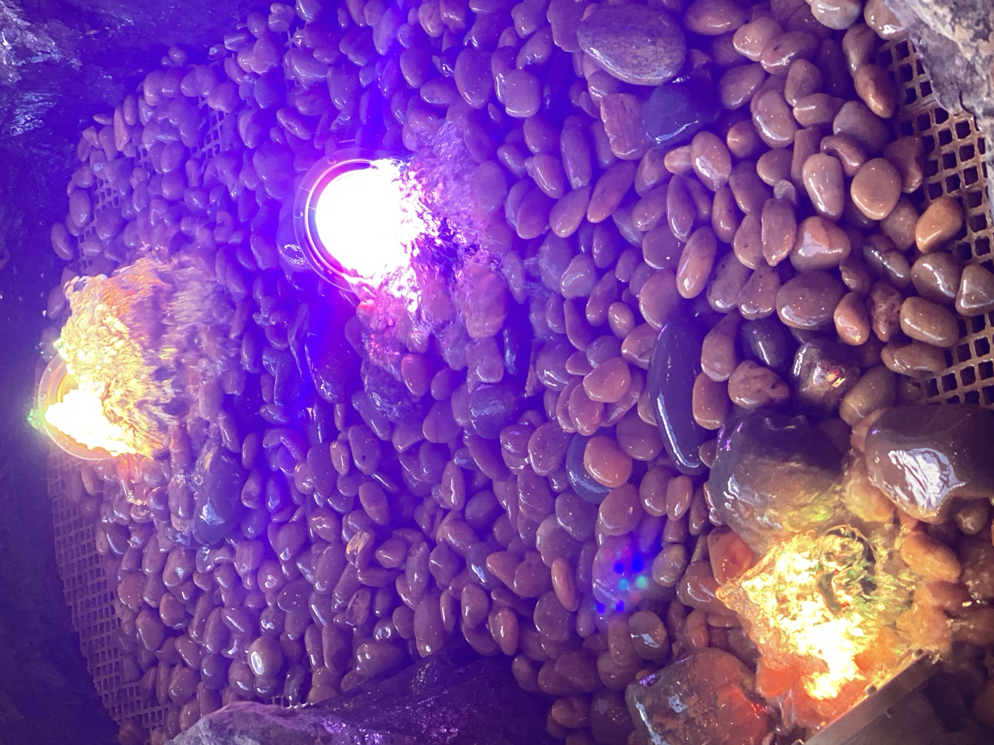 The image shows a close-up view of a decorative fountain filled with smooth, rounded pebbles. Two illuminated water features are visible. One, on the left, emits a bright orange and yellow light, suggesting a warm glow. The other, on the right, is illuminated by a bright pink and purple light, with water cascading around it. The pebbles are predominantly in shades of brown and grey, with some darker, wet-looking stones interspersed. A mesh grate is visible beneath the pebbles in some areas, suggesting the structure of the fountain. The overall lighting is dark, with the purple hue from the fountain lights creating an ambient glow over the scene. There are no people visible in the image. The scene appears to be an indoor or partially enclosed decorative water feature, likely a part of a public space or private garden. There is no information in the image to specifically place it in Novokuznetsk, Russia.