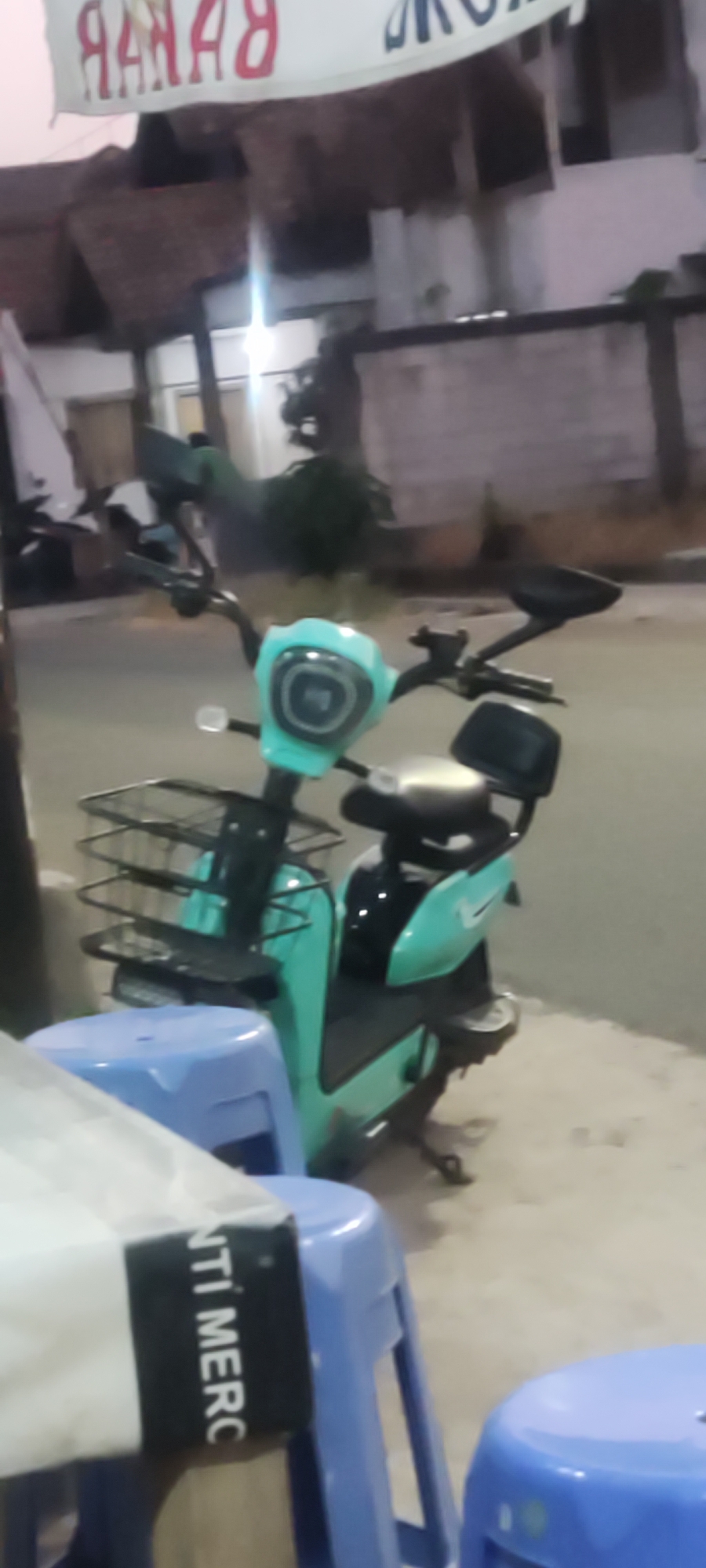 The image shows a turquoise electric scooter parked outdoors, possibly on a sidewalk or street. The scooter has a basket attached to the front and a black seat with a backrest. In the foreground, a portion of a white banner with red lettering, which appears to be reversed and could read "RANAH" or something similar, is visible. To the left and right of the scooter, two blue plastic stools are partially visible. The background suggests an urban environment with buildings and some vegetation. The lighting appears to be dim, indicating it might be dusk or nighttime. The location is specified as Getrakmoyan, Indonesia.