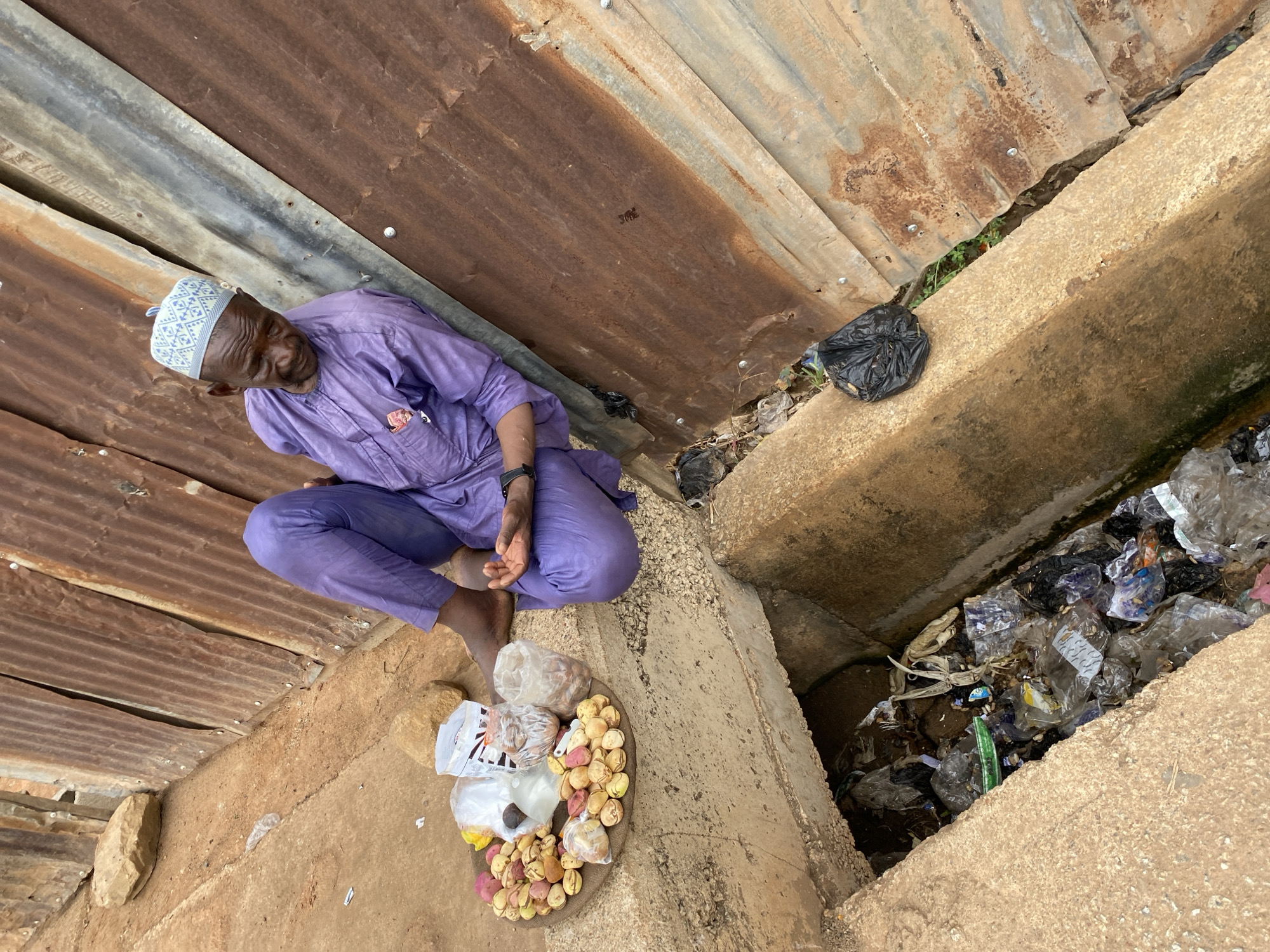 A man in a purple outfit and patterned cap sits cross-legged on a dirt ground next to a corrugated metal roof. In front of him, on a round tray, are various items including loose nuts and plastic bags of goods. To the right of the man is a concrete drainage ditch filled with trash. The scene appears to be in Bukuru, Nigeria.