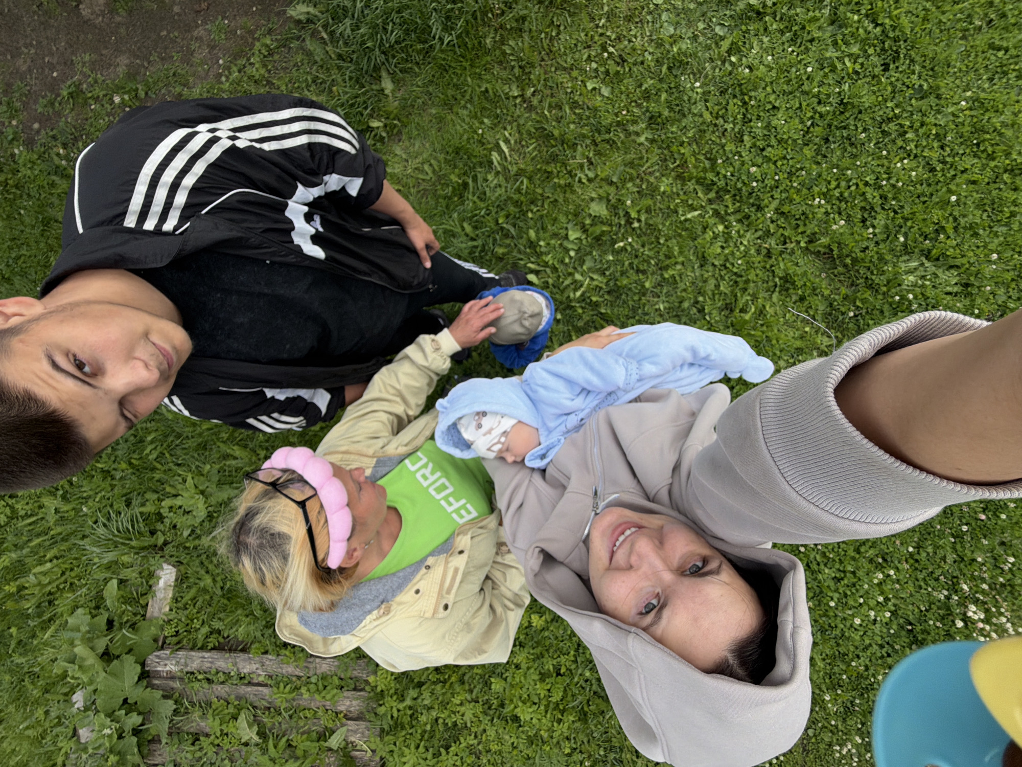 This media file depicts three people and a baby lying on a grassy area, likely outdoors in Andreyevo, Russia. The perspective is from above, looking down on the subjects.
In the upper left of the image is a young person wearing a black Adidas jacket with white stripes. Next to them, to the right, is another person wearing a light-colored jacket over a bright green t-shirt. They have blonde hair, glasses, and a pink headband. The green t-shirt has visible white text that appears to read "REFORC". This individual is also holding a baby wrapped in a blue blanket, with the baby wearing a patterned hat. In the lower right of the image is a person wearing a grey hooded sweatshirt, looking up and smiling at the camera.
The scene is set outdoors on a lawn with scattered small white flowers and some exposed wooden planks. The lighting suggests it is daytime, likely overcast given the soft shadows. The overall mood appears relaxed and familial. The presence of a baby suggests a family gathering or outing.