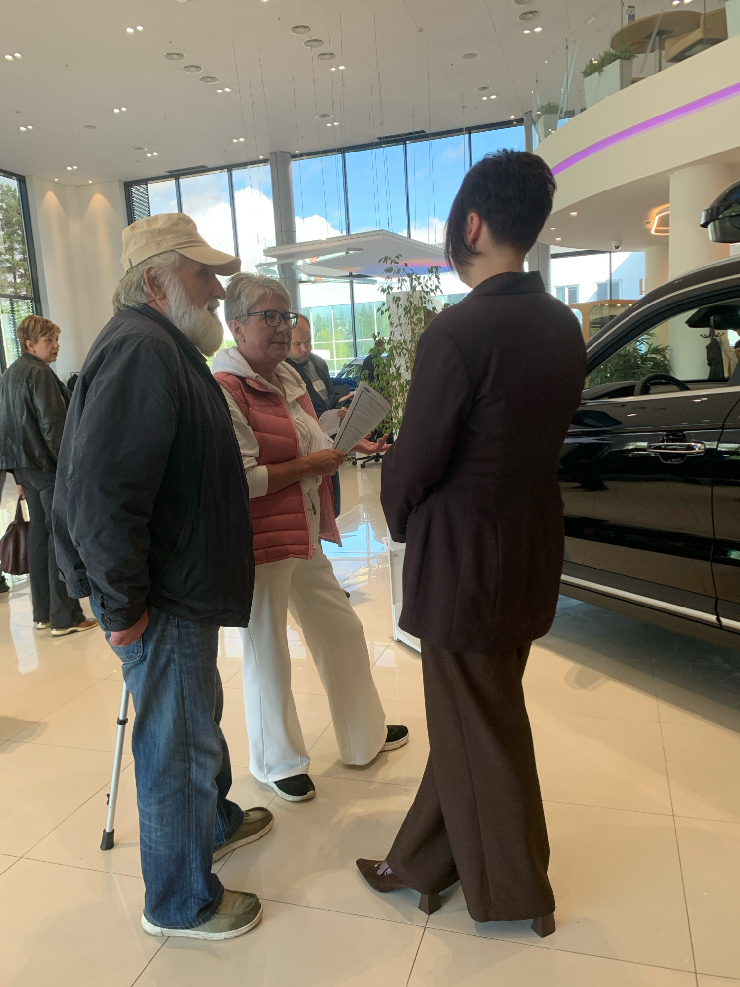 In a well-lit showroom, possibly a car dealership, located in Minsk, Belarus, four individuals are engaged in a conversation near a black SUV. On the left, an older man with a beard and a beige cap leans on a crutch, wearing a dark jacket and blue jeans. Beside him, a woman with short grey hair and glasses wears a pink vest over a white top and white pants. A third person, facing away from the camera, is dressed in a formal brown suit. To the far left, another woman in a black jacket is partially visible. The environment features large windows with a view of trees and the sky, polished tile flooring, and modern lighting fixtures.