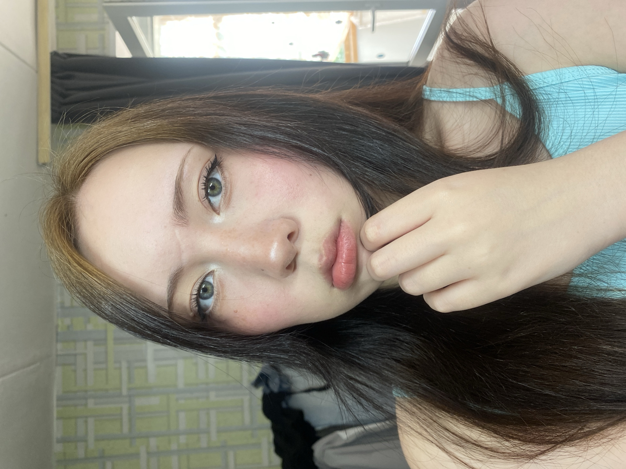 A young woman is posing for a close-up selfie. She has long, dark hair with blonde highlights, light skin, and light eyes with black eyeliner. She is wearing a light blue top. Her right hand is positioned near her face. The background includes a window with black curtains and a patterned wall. The location is unknown.