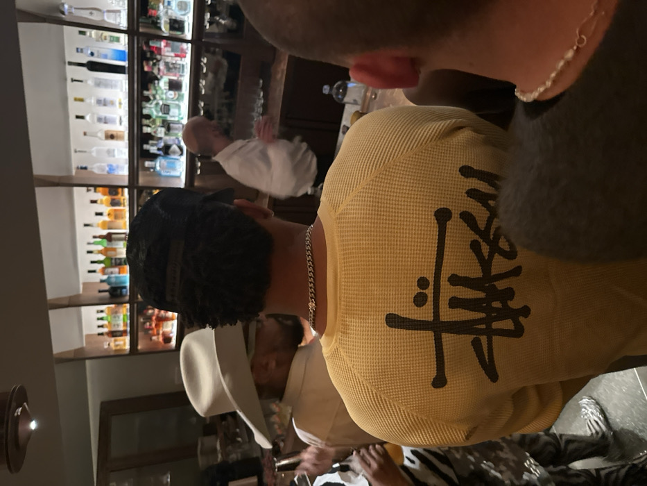 A group gathers in a dimly lit room, featuring a bar with multiple shelves filled with bottles of alcohol. A person in a yellow waffle-knit shirt emblazoned with a black logo faces away from the camera. A bald man wearing a white shirt is visible in the background near the bar. A person wearing a cowboy hat stands nearby. Another individual with a silver chain and a light grey shirt is also present.