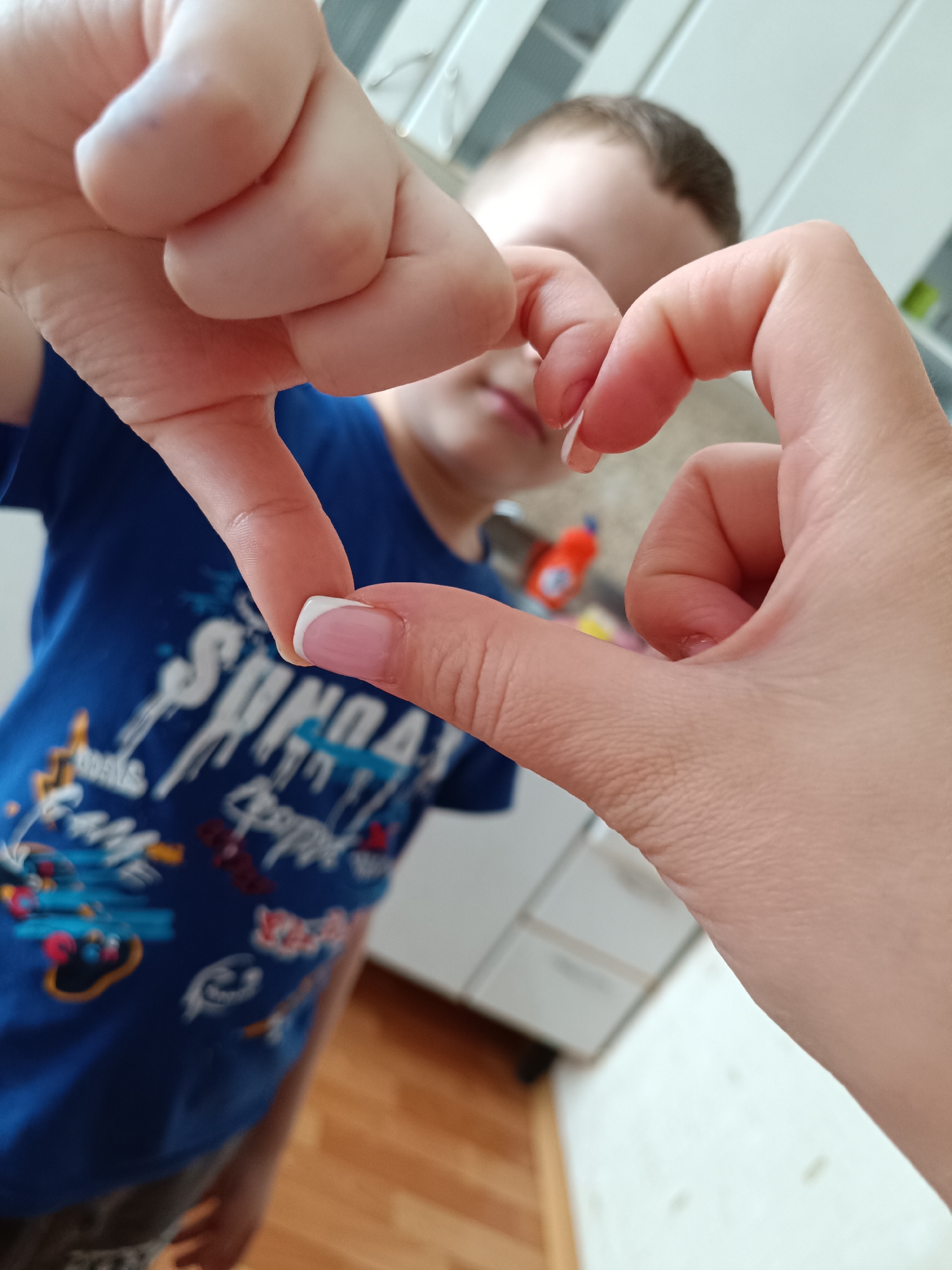 A person with fair skin and French-manicured nails forms a heart shape with their hands. A child wearing a blue t-shirt stands slightly out of focus in the background. The location appears to be indoors, potentially a home, judging by the kitchen cabinetry in the background and the wooden flooring. The city of Tobolsk, Russia, cannot be confirmed from the visual evidence alone.