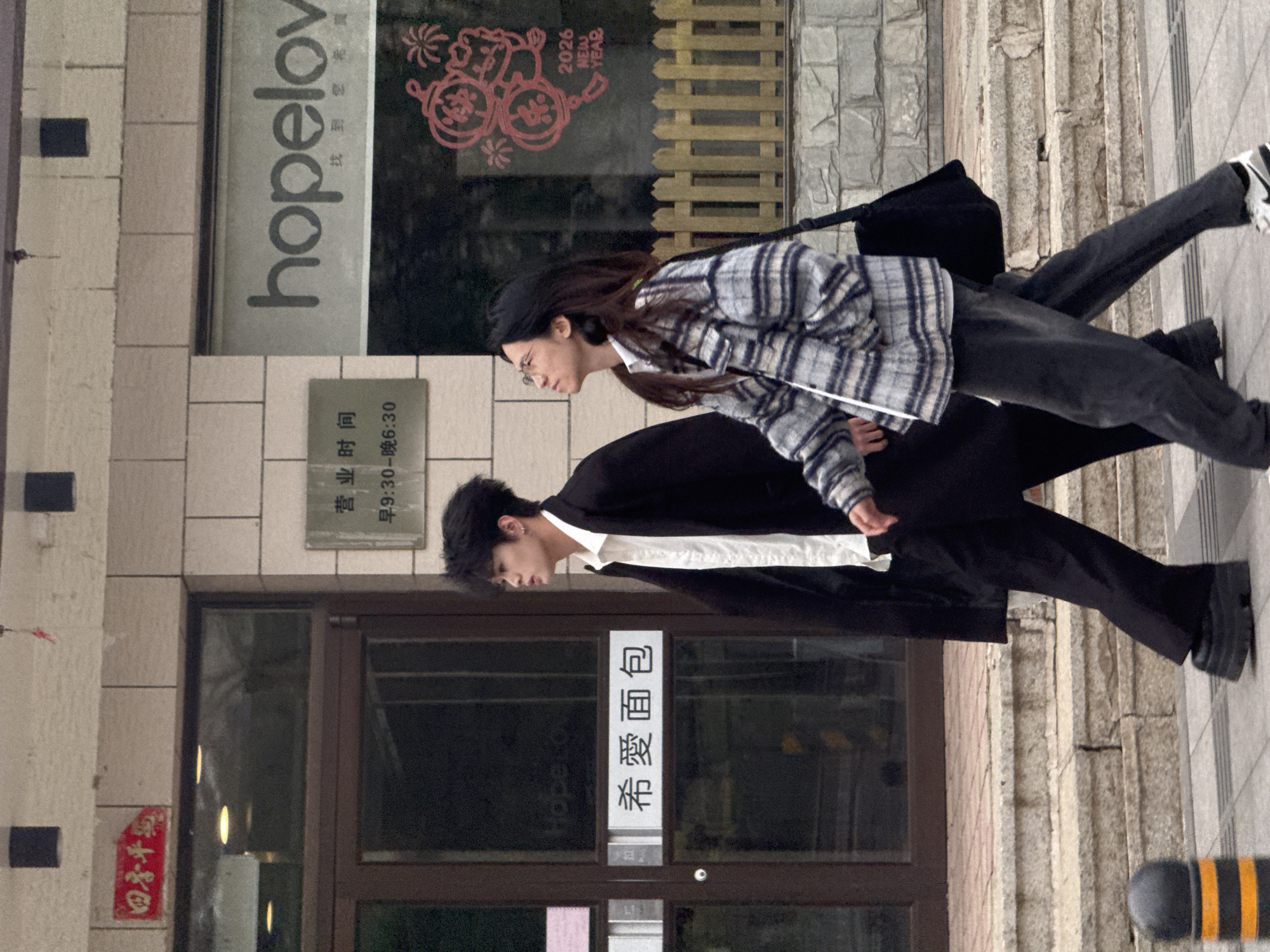The image shows two people standing on a sidewalk in front of a building. The person in the foreground is wearing a black blazer over a white shirt and black pants. The person in the background is wearing a grey and white plaid shirt, dark jeans, and a black beanie. The building behind them has a glass door with Chinese characters that read "Xi Ai Mianbao" (希愛面包), which translates to "Hope Love Bread." Above the door is a sign indicating business hours, from 9:30 AM to 6:30 PM. Above that, another sign says "hopelov," and a New Year decoration shows the year 2026. Based on the signs, the location is a bakery called "Hope Love" and likely located in Shenyang City, China.