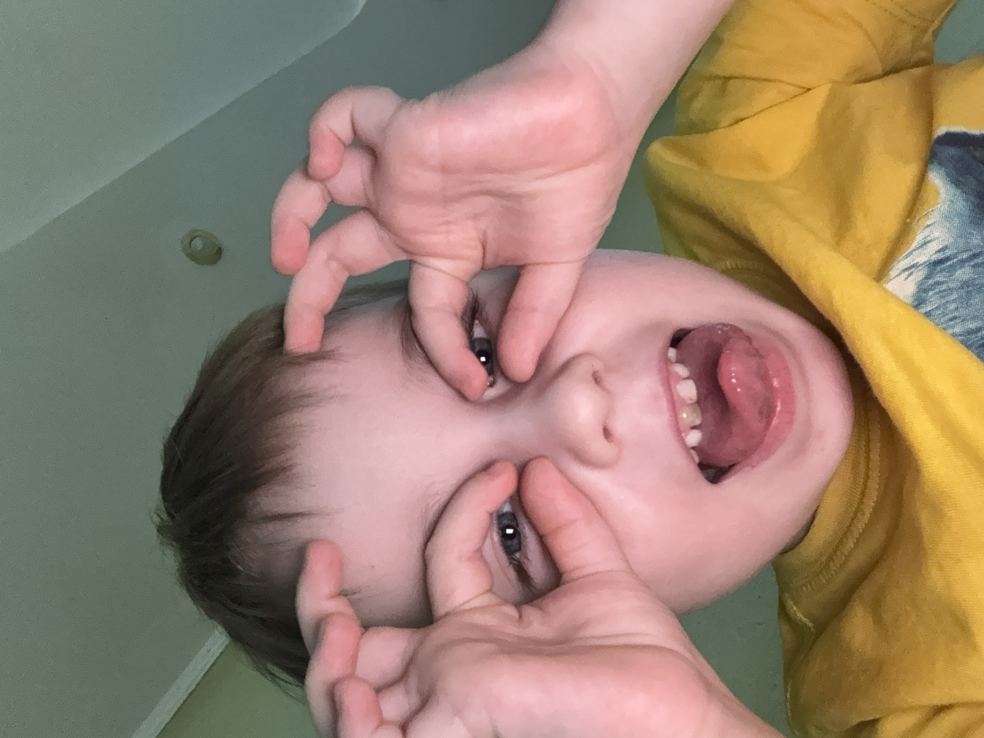A young child, viewed from a low angle, is making a face. The child is wearing a yellow shirt and pulling their eyelids apart with their fingers while sticking their tongue out. The background appears to be a pale green wall and ceiling with a recessed light fixture. The location cannot be determined to be Chaikovsky, Russia from the available visual information.