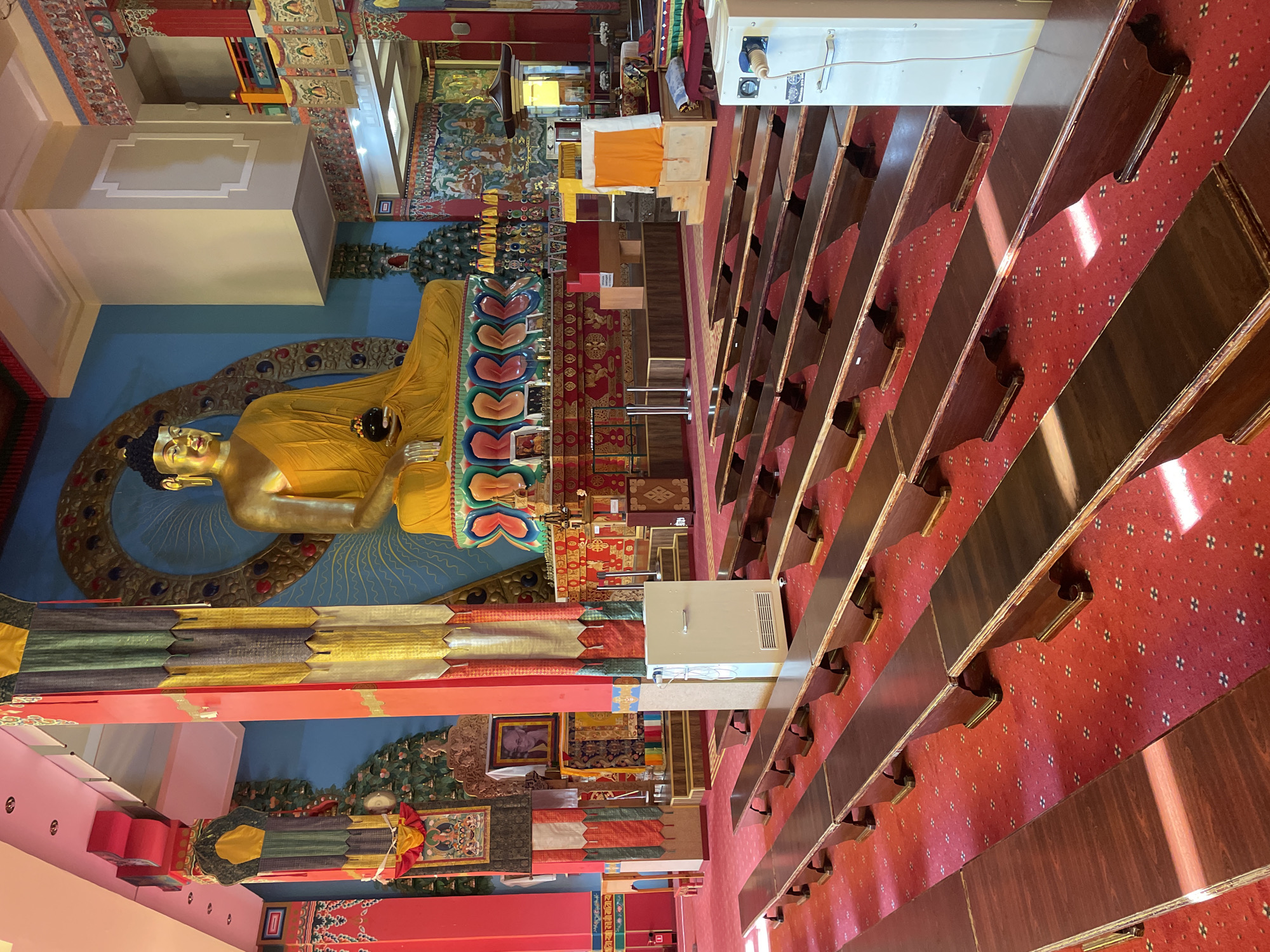 This is an indoor photograph of the interior of the Golden Temple of Elista, Russia. The primary subject is a large, golden Buddha statue seated on a colorful pedestal. The statue faces forward, and its hands are resting in its lap, holding a dark object. The Buddha is adorned in yellow robes.
The foreground is dominated by rows of dark wooden benches, arranged in parallel lines on a bright red carpet. The carpet has a repeating small, dark pattern. The benches are simple in design, with curved wooden legs supporting the seating surface. There are two large, white, rectangular air conditioning units visible, one in the middle ground to the left and another in the middle ground to the right.
The background features a vibrant blue wall behind the Buddha statue, decorated with a large, circular, ornate golden design. To the sides of the Buddha are tall, red, decorative pillars adorned with colorful fabric banners in shades of yellow, grey, and red, hanging vertically. On the walls in the background, there are various religious paintings and images, some framed and some part of larger murals. Further in the background, there are more decorative elements, including what appear to be smaller statues and religious artifacts on shelves and altars. The ceiling is a light color, with visible lighting fixtures. The overall impression is a richly decorated and serene religious space.