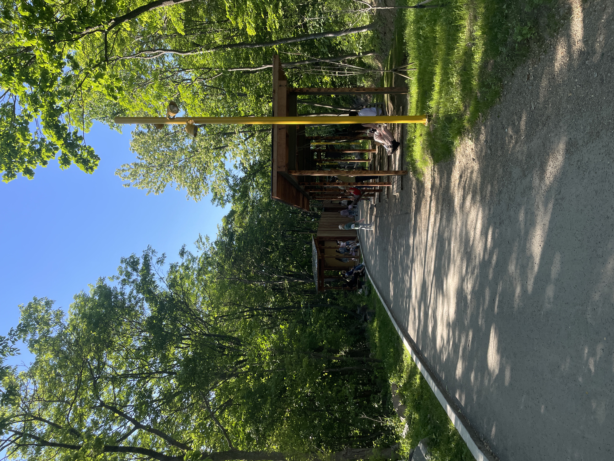 A daytime, eye-level shot depicts a park setting in Vladivostok, Russia.
Two wooden structures with roofs, appearing to be shelters or bus stops, stand amidst lush green trees. People occupy the seating areas within the structures. A yellow horizontal bar with lights is above the first structure.
A paved pathway runs alongside the wooden structures, with grass bordering the side. Sunlight casts shadows of trees onto the path. The sky is blue and visible through gaps in the tree canopy.